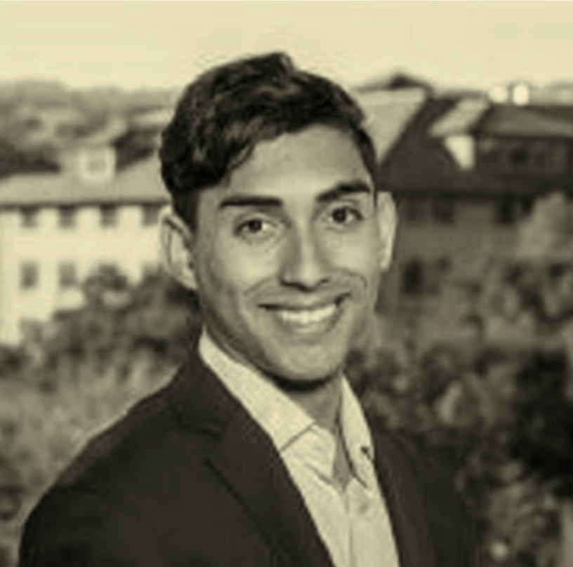 A portrait of a young man with dark hair, wearing a suit and light-colored shirt, smiling outdoors with buildings in the background.
