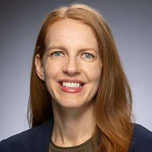 A woman with long red hair, blue eyes, and fair skin, smiling and wearing a dark blazer.
