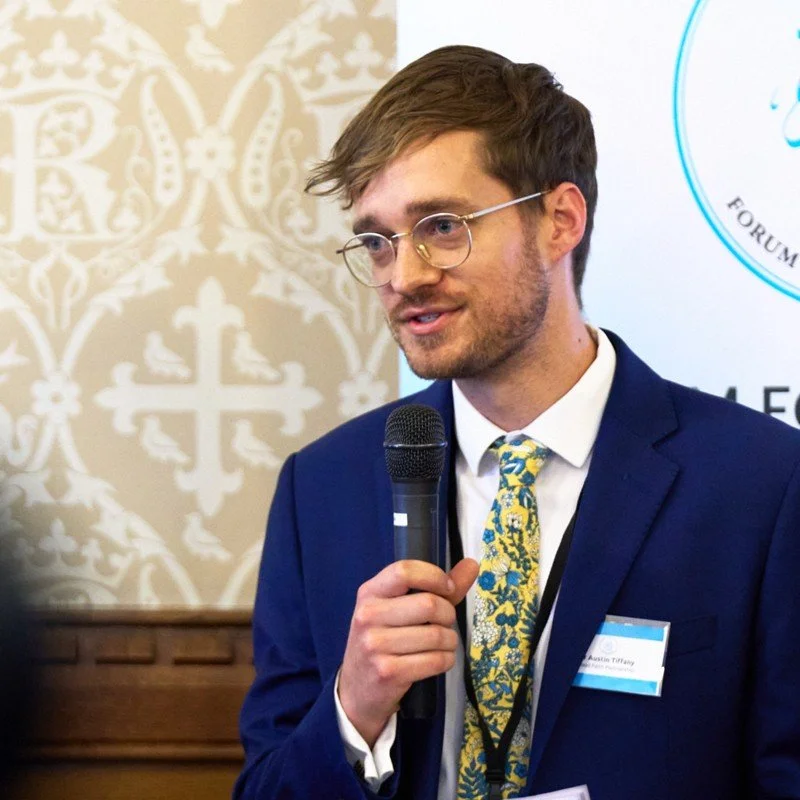 A man in a blue suit holding a microphone, speaking at an indoor event with a patterned wall and banner in the background.
