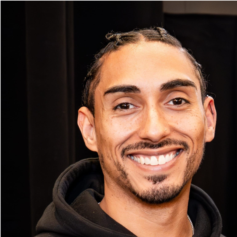 Close-up of a smiling man with dark hair, braid detail, goatee, wearing a black hoodie, against a dark background.