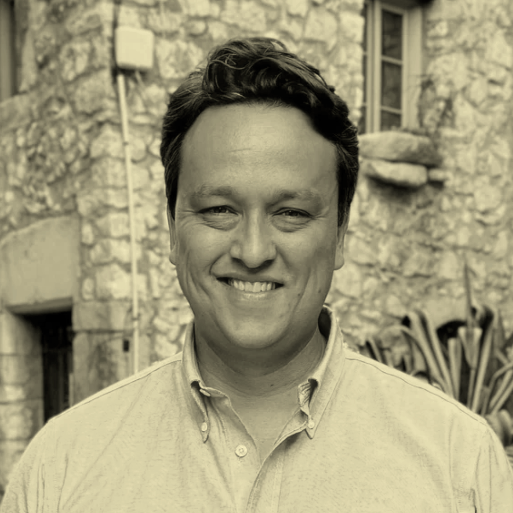A smiling man with dark hair, wearing a collared shirt, standing outdoors in front of a stone building.