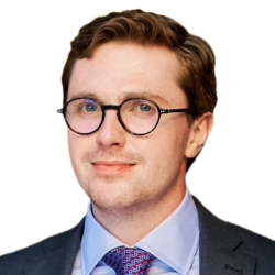 Portrait of a man with light brown hair, glasses, wearing a dark suit, light blue shirt, and patterned tie, against a white background.