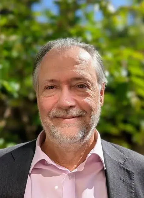 A middle-aged man with gray hair, beard, and mustache, wearing a pink shirt and gray blazer, smiling outdoors with a blurred green foliage background.