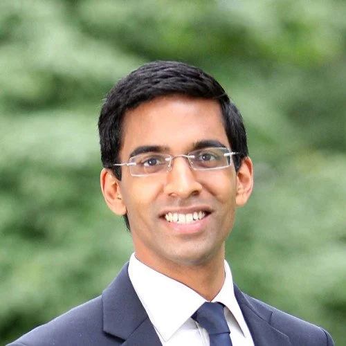 Professional portrait of a man in a suit with glasses, smiling outdoors with a blurred green background.