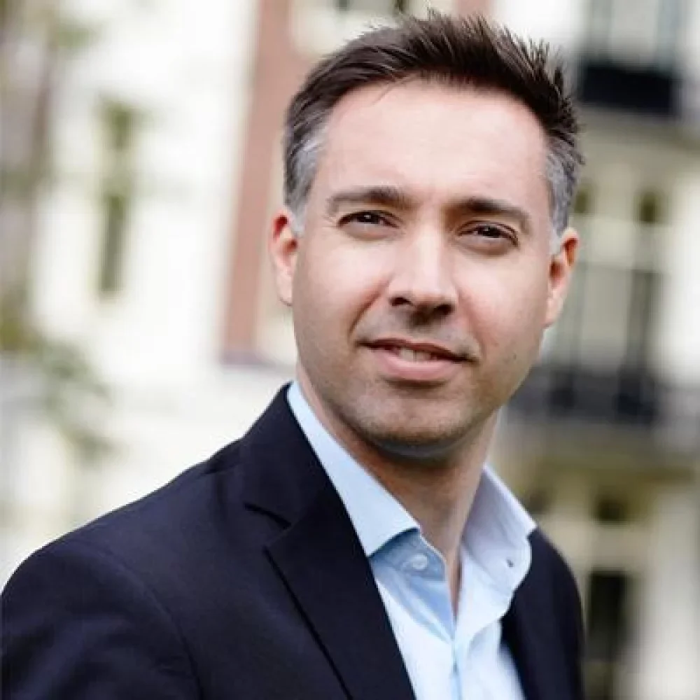 A man with short brown hair wearing a dark suit jacket and light blue dress shirt outdoors.