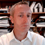 A man with short gray hair and a slightly serious expression, wearing a black suit jacket and a white shirt, against a plain white background.