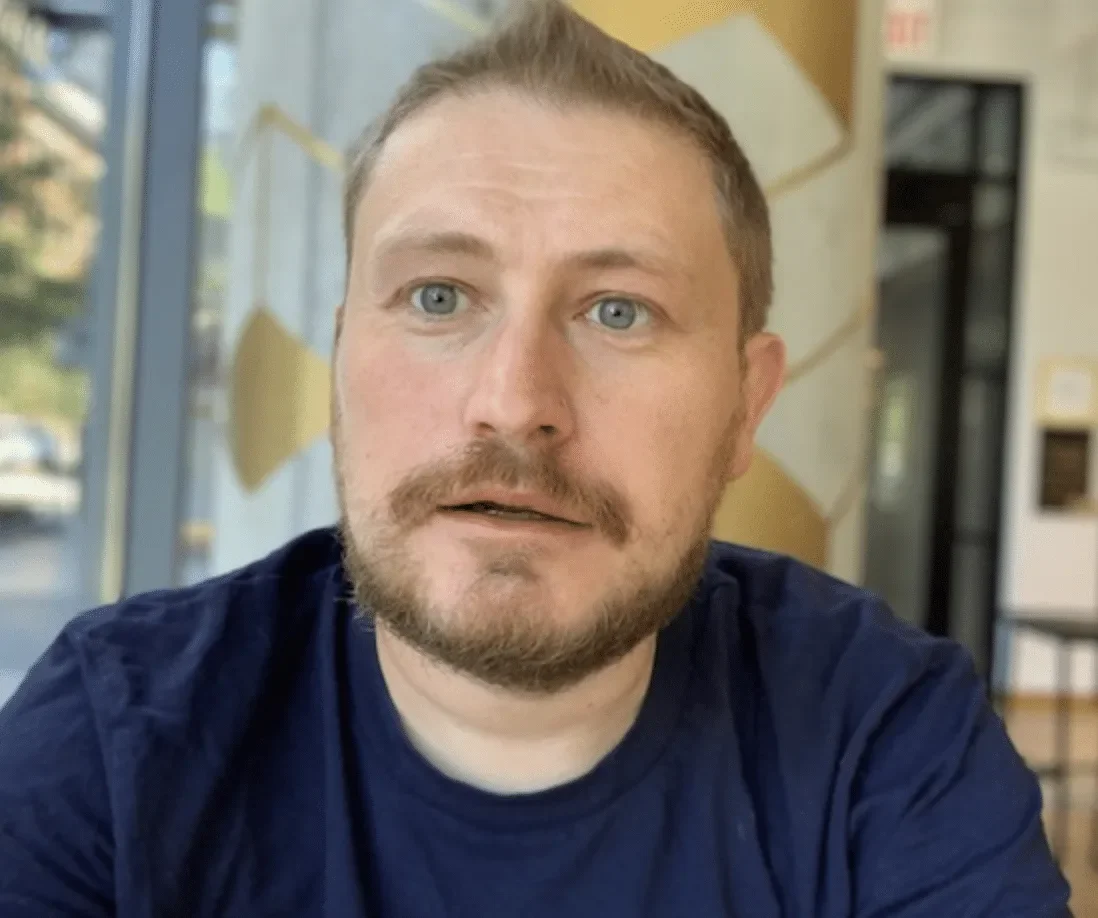 A man with blue eyes, light brown hair, and a beard wearing a navy blue shirt, sitting indoors, with a patterned wall and window in the background.
