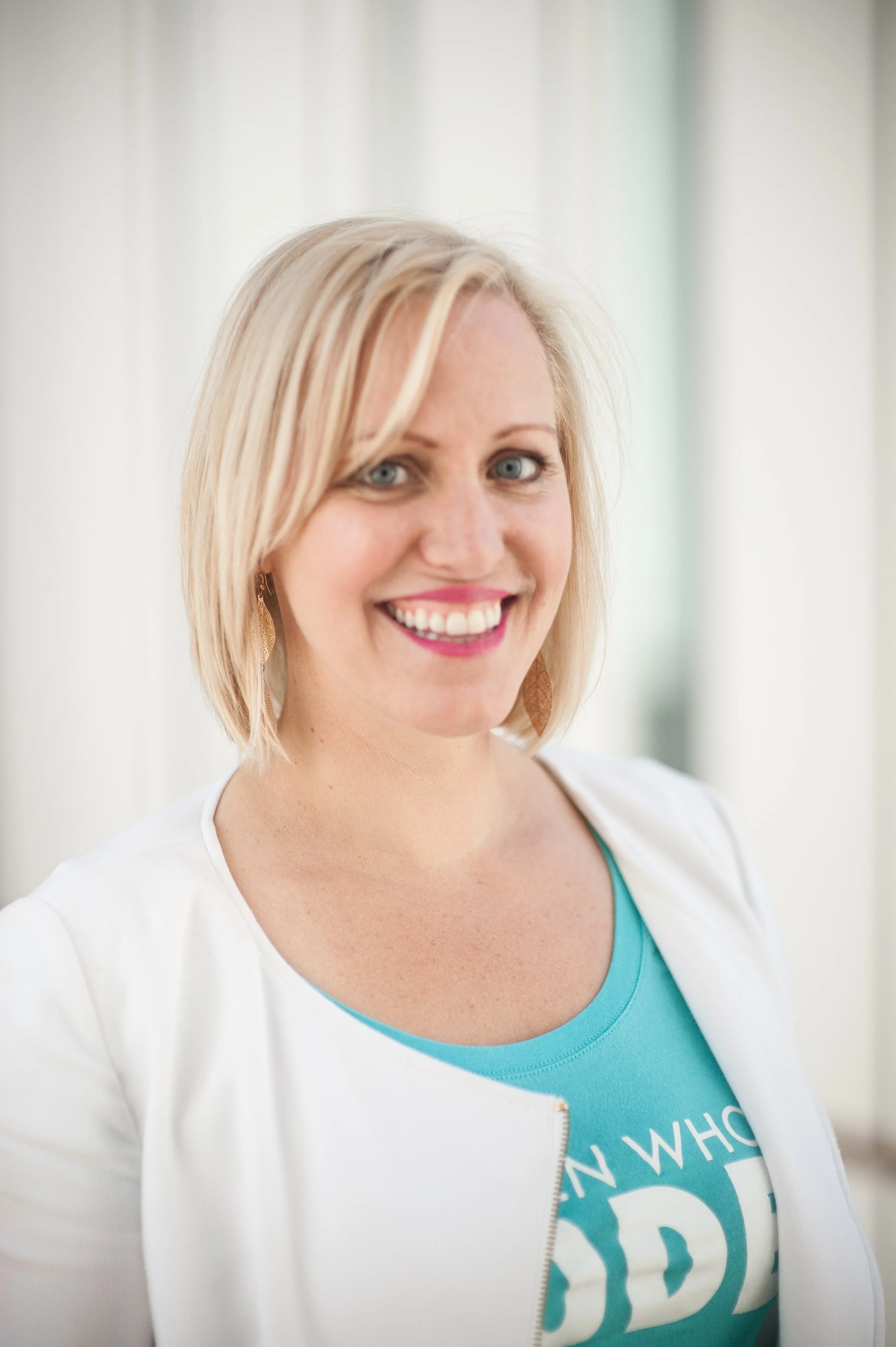 A woman with blonde hair, blue eyes, and a bright smile, wearing a white jacket over a teal shirt with partial text visible. She is standing indoors with a light, blurred background.