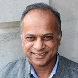 Close-up of smiling middle-aged man with short hair, wearing a blue shirt and gray blazer, standing outdoors in front of concrete wall.
