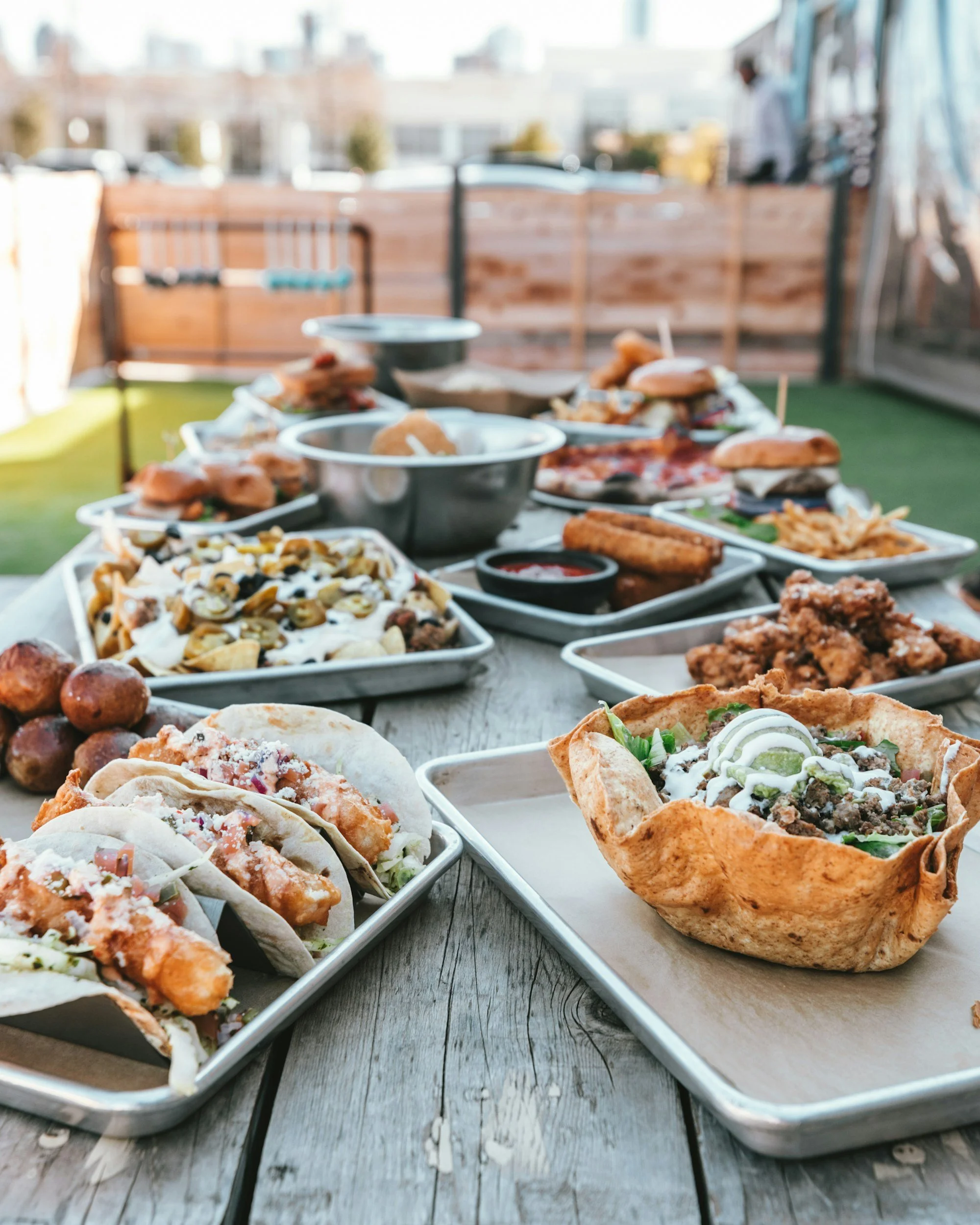 Outdoor table filled with various dishes including tacos, fried chicken, pasta salad, wings, burgers, and sides at a backyard gathering.