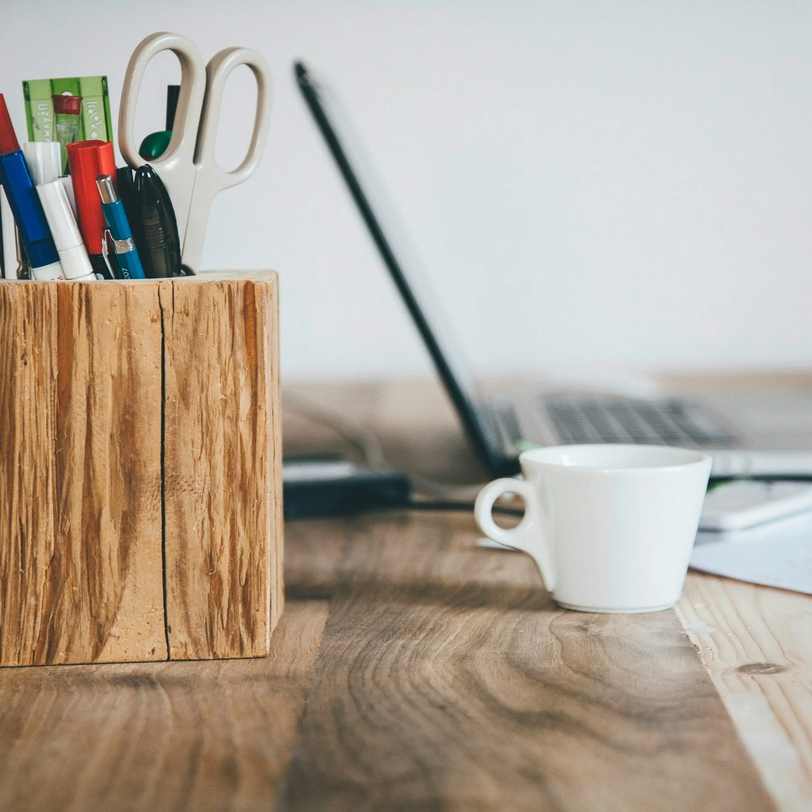 A wooden desk with a white mug, a laptop, and a wooden container holding pens, markers, and scissors.