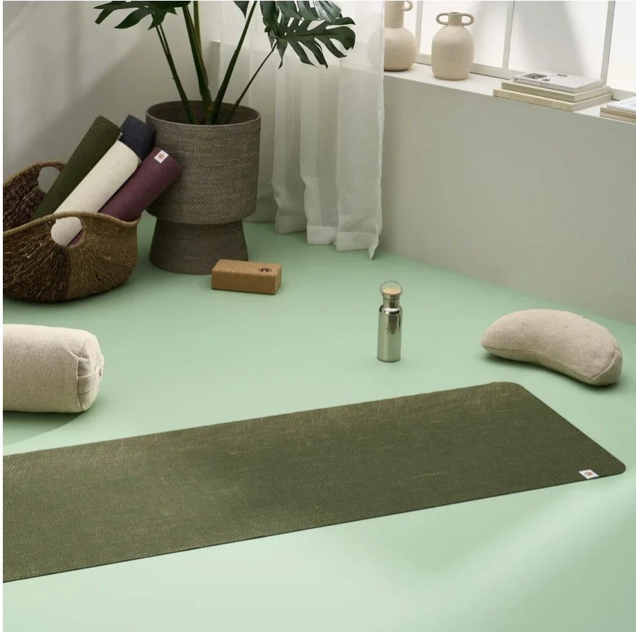 Yoga mat, water bottle, meditation pillow, rolled towel, plant, two vases, books, curtains, and window in a bright, minimalist room.