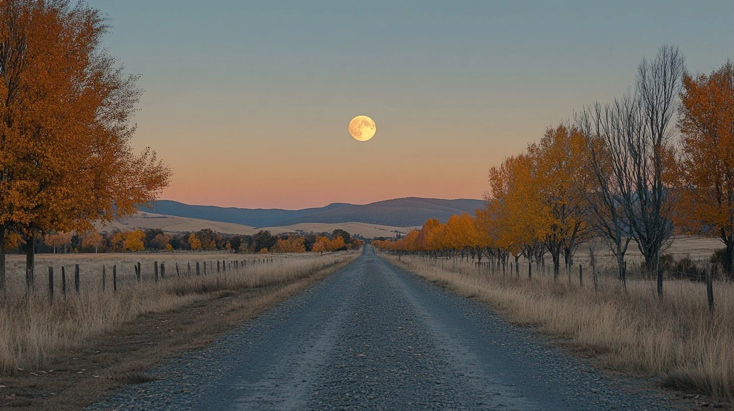 A rural dirt road lined with autumn trees under a clear sky, with the full moon visible near the horizon.