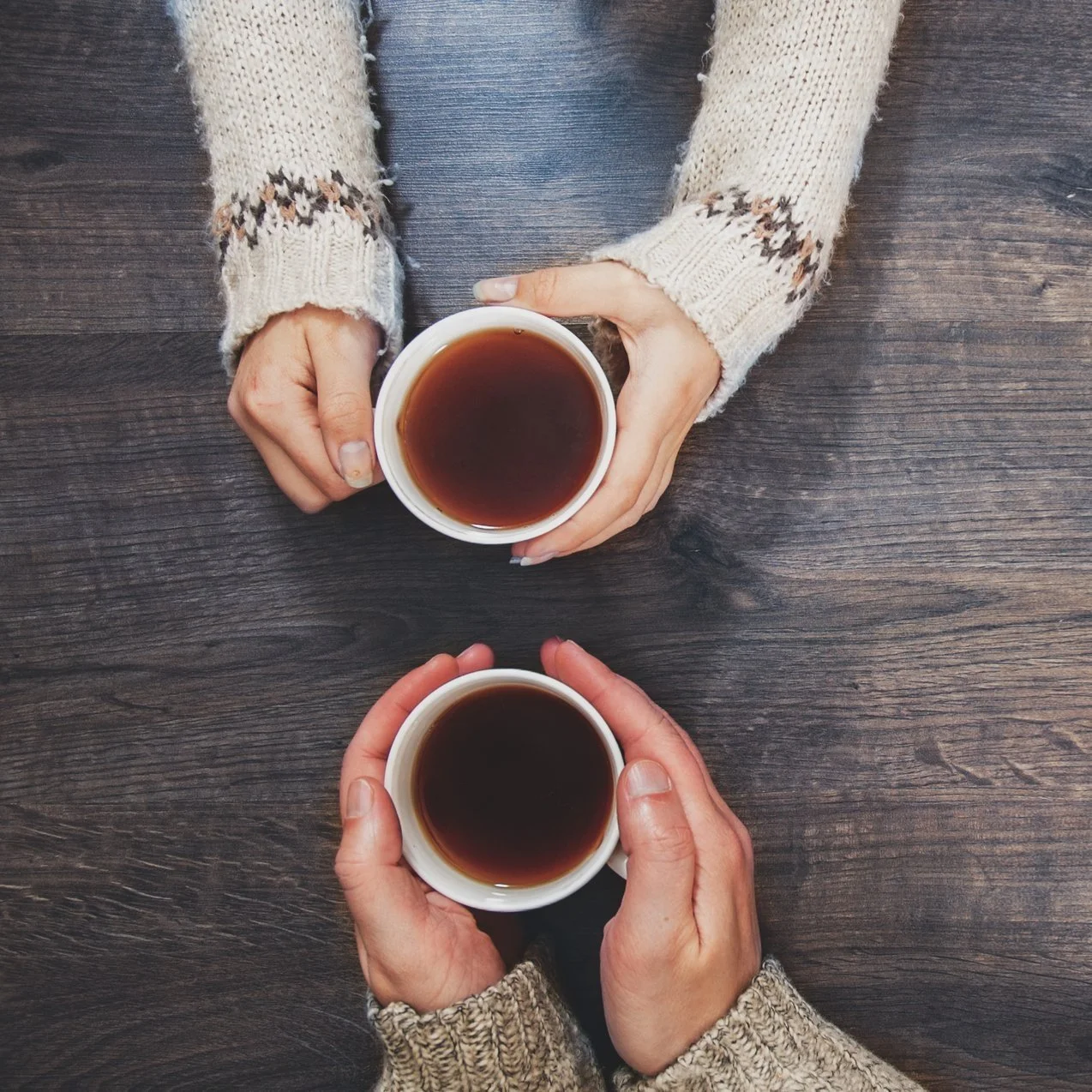 Two people holding white mugs filled with black coffee over a dark wooden table, viewed from above.
