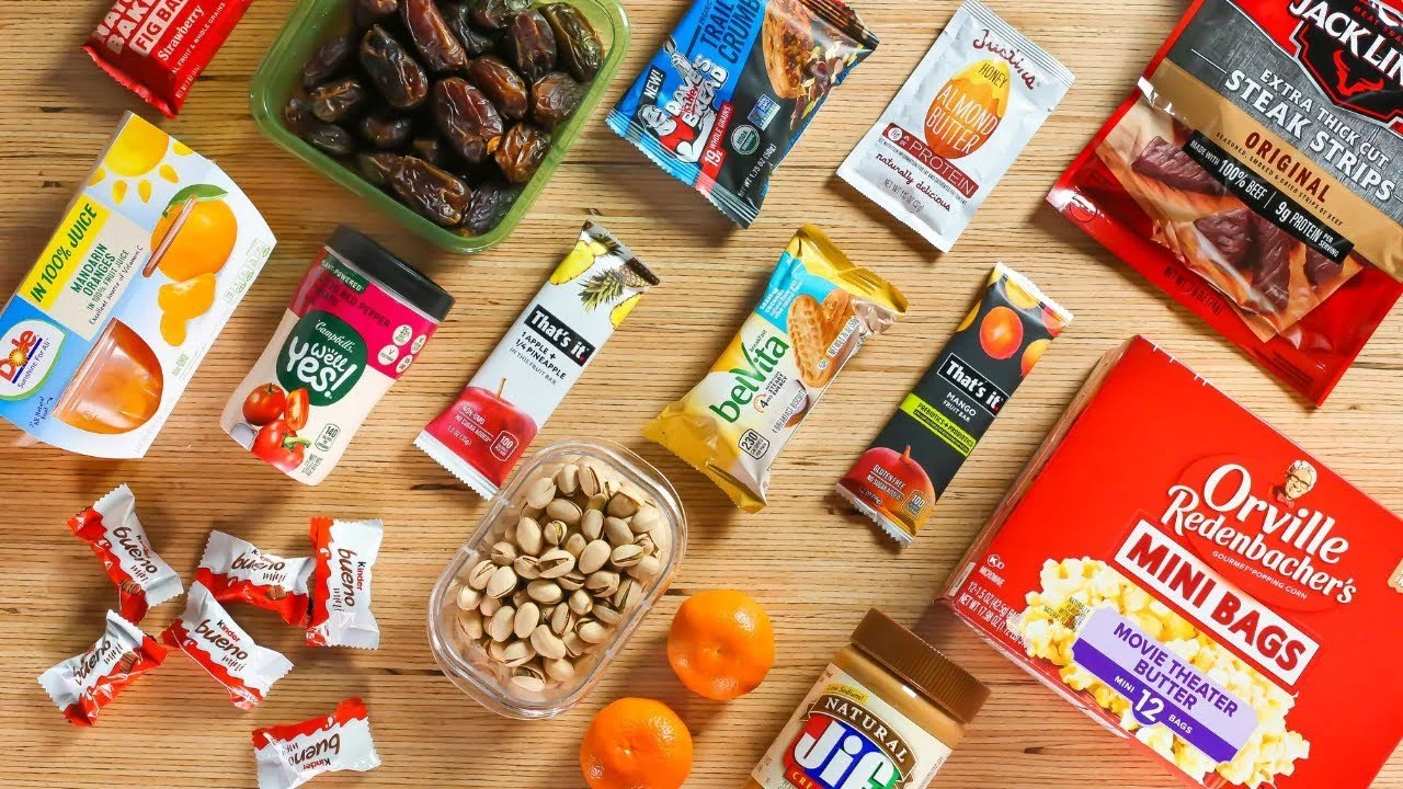 A variety of snack foods and beverages spread out on a wooden surface, including dates, pistachios, mandarin oranges, chocolate-covered candies, snack bars, juice boxes, mini bags of popcorn, and peanut butter.