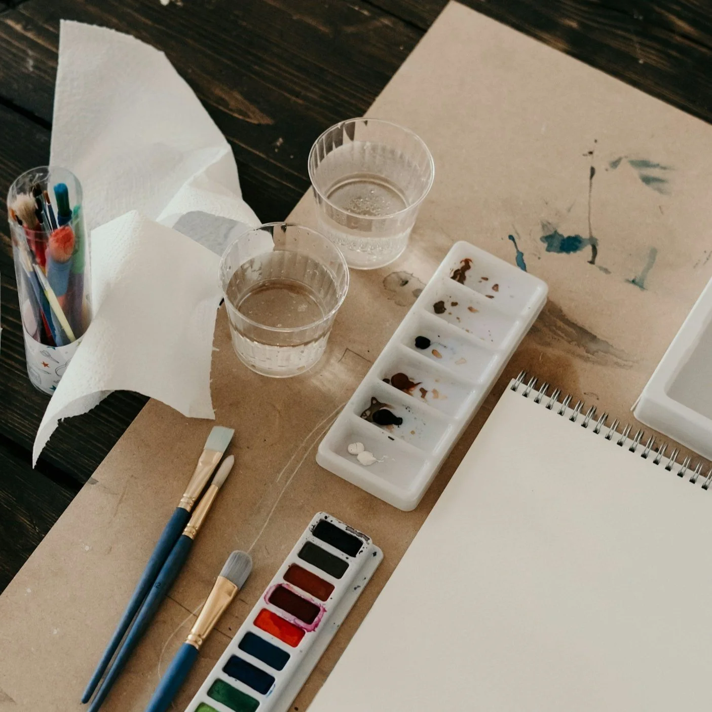 A watercolor painting setup with brushes, a palette with mixed paint, two glasses of water, and a blank sketchbook on a brown paper sheet on a dark wooden table.
