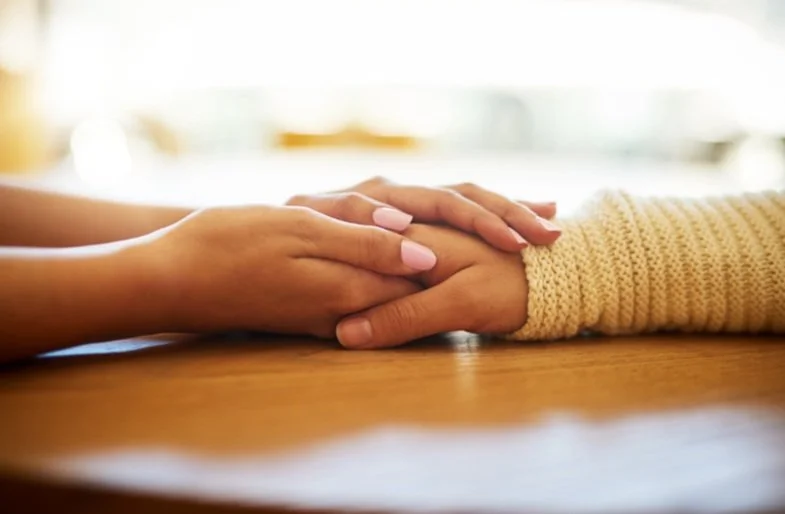 Two people gently holding hands with one person's hand resting on top of the other's on a wooden table.