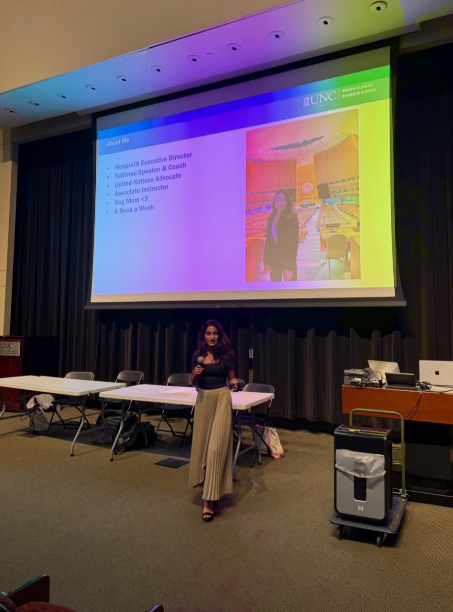 A woman stands in front of a large projection screen in a conference room, speaking to an audience. The screen displays a presentation slide with her photo, a colorful auditorium, and text about her roles and interests. The room has tables and chairs, and some equipment like a microphone, laptop, and trash bin are visible.