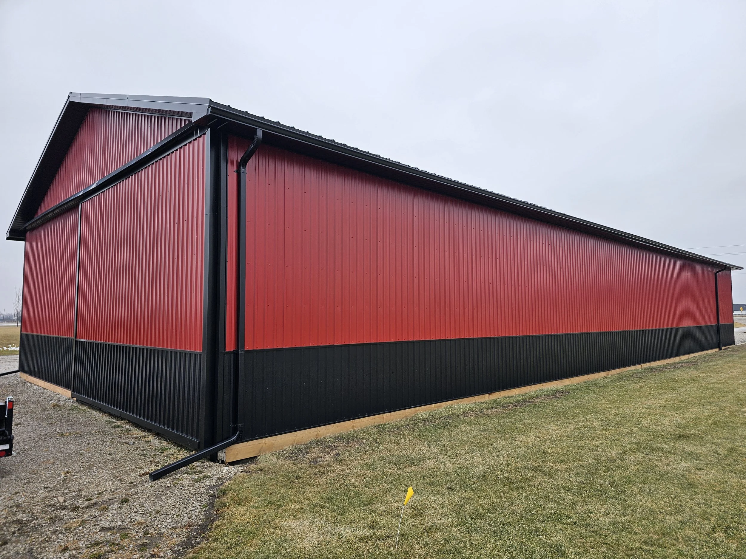 Large red metal building with black trim and a sloped roof, situated on a grassy area under a cloudy sky.
