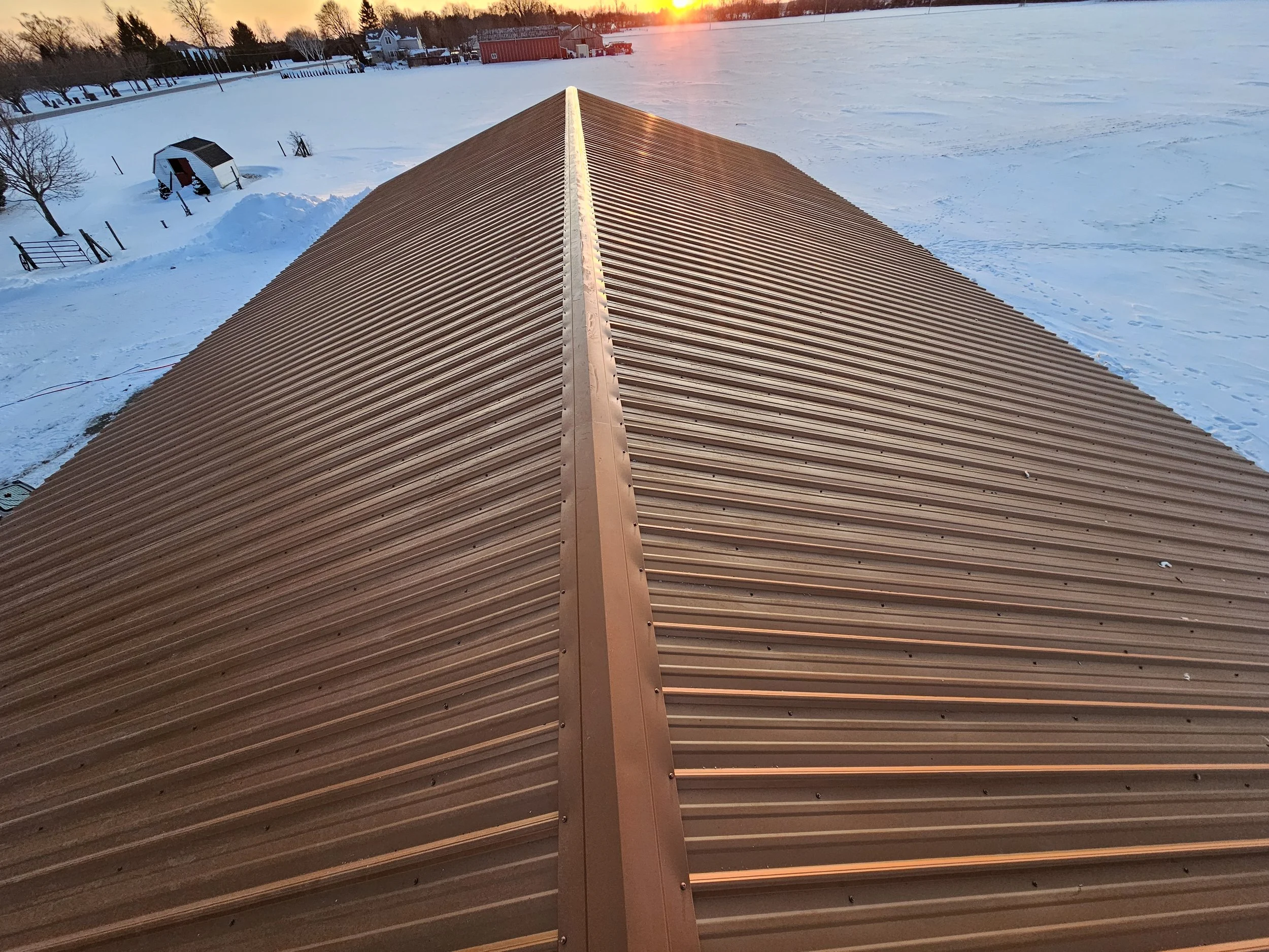 Looking over the roof of a barn or shed with brown metal roofing, with a snowy landscape and the setting sun in the background.
