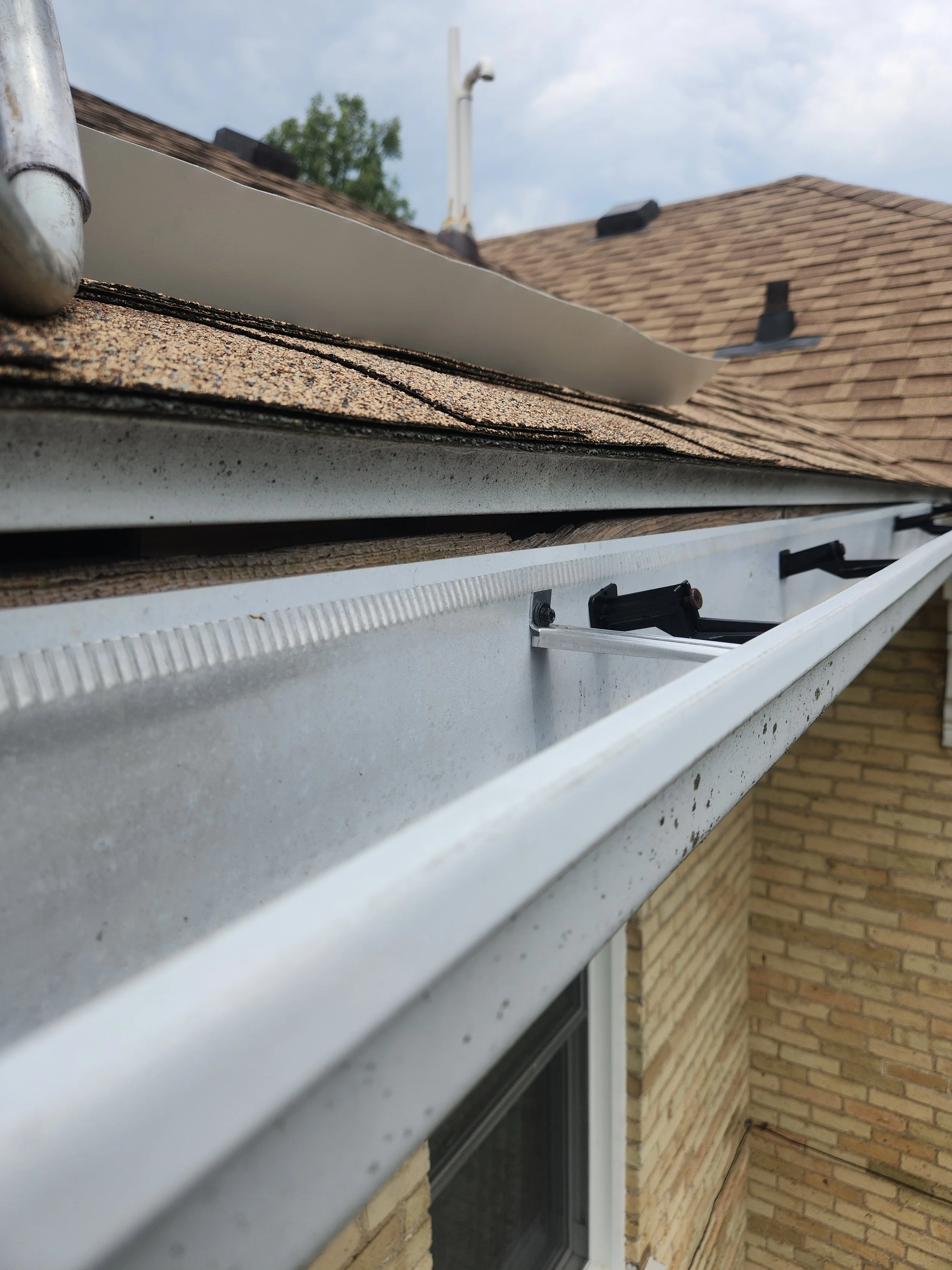 Close-up of an eavestrough (gutter) on a residential house, showing a section of the gutter, part of the roof with shingles, and a brick wall below.