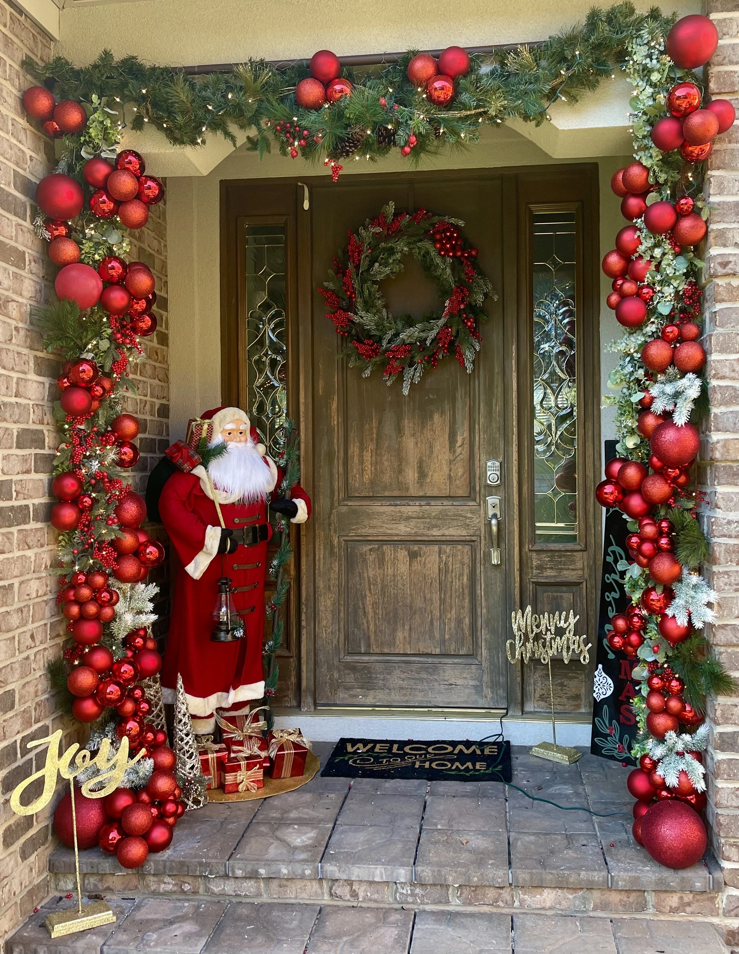 Decorated Christmas front porch with a large green and red ornament garland framing a wooden door, a Santa Claus figure, wrapped presents, and festive signs with holiday greetings.