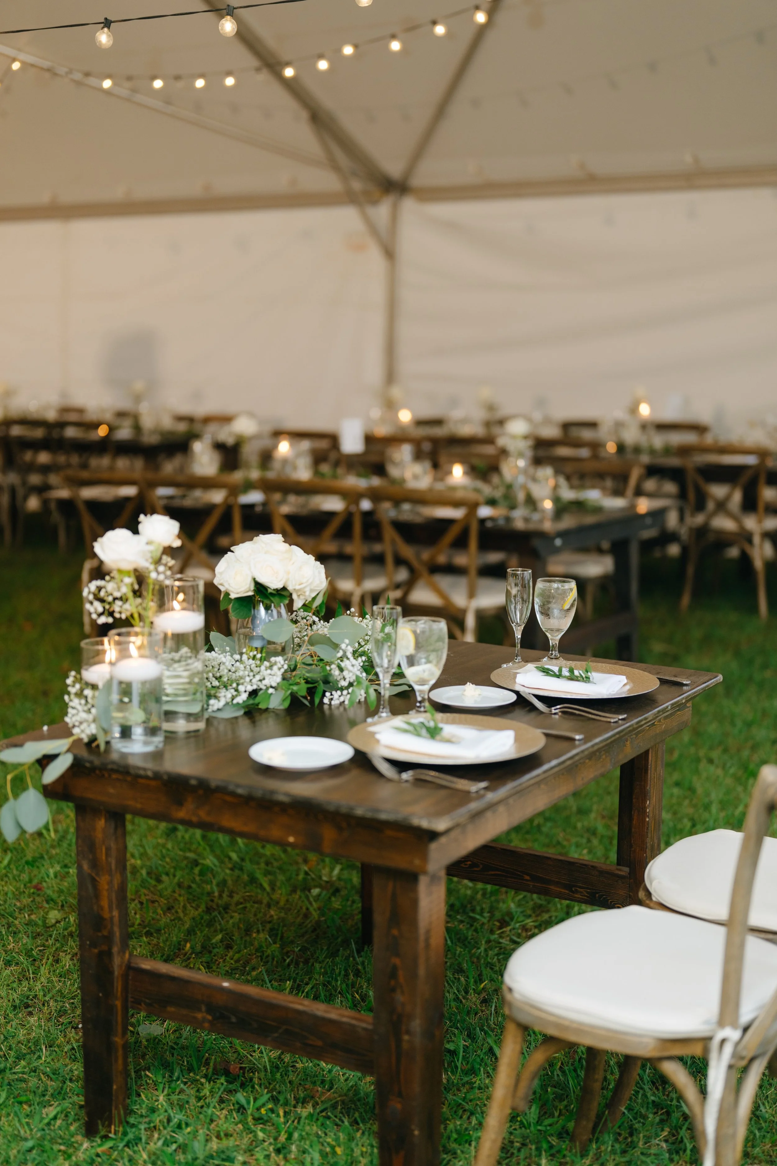 A stunning wedding reception, complete with wooden tables, greenery centerpieces, charger, white dishes, and farmhouse-style chairs; styled by Laura at Design & Gather.