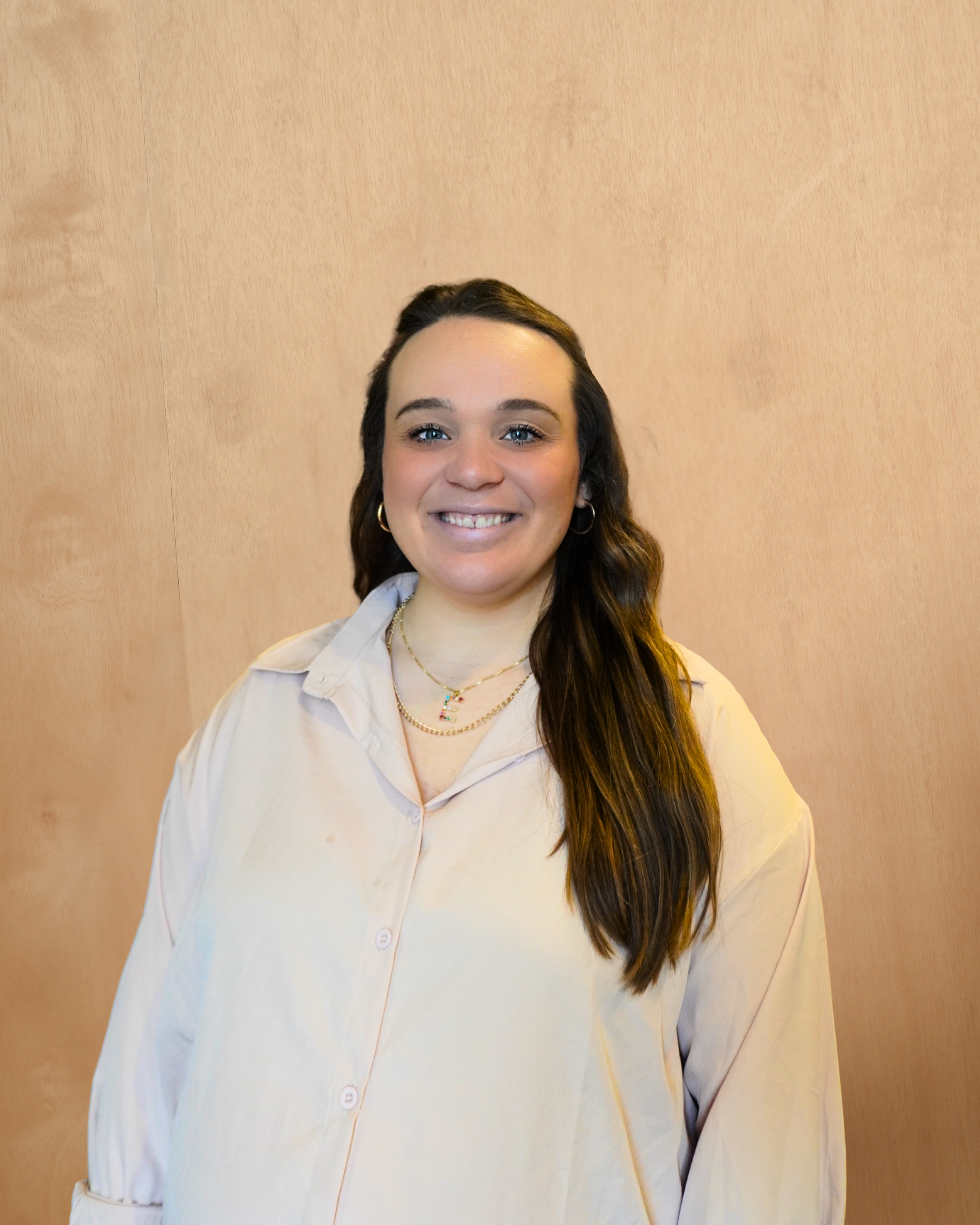 Smiling woman with long brown hair, wearing a light beige button-up shirt and layered necklaces, standing against a wooden background.