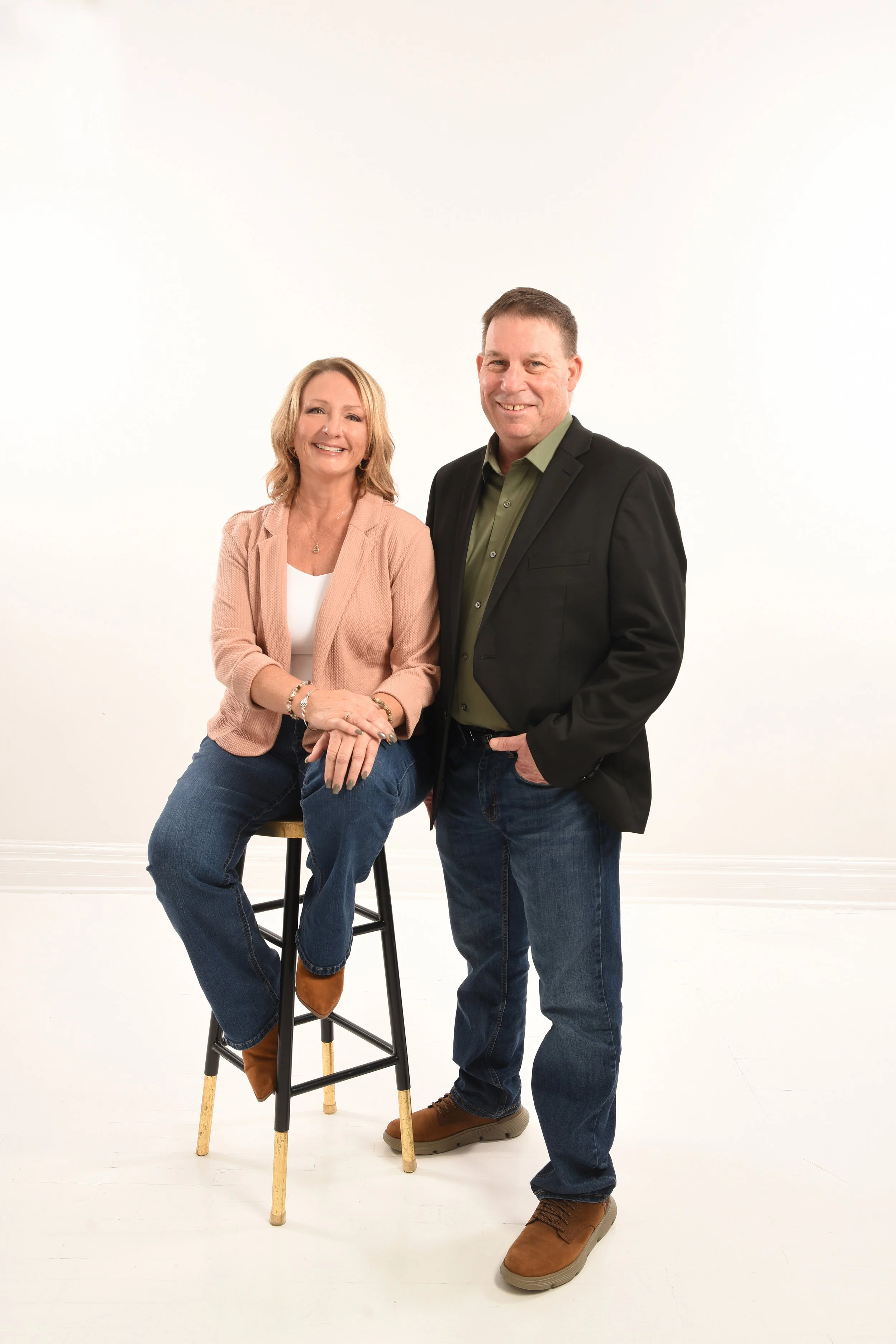 A woman sitting on a stool with a man standing beside her in a white studio setting. The woman is wearing a light pink blazer, white top, and jeans. The man is dressed in a black blazer, green shirt, and jeans.