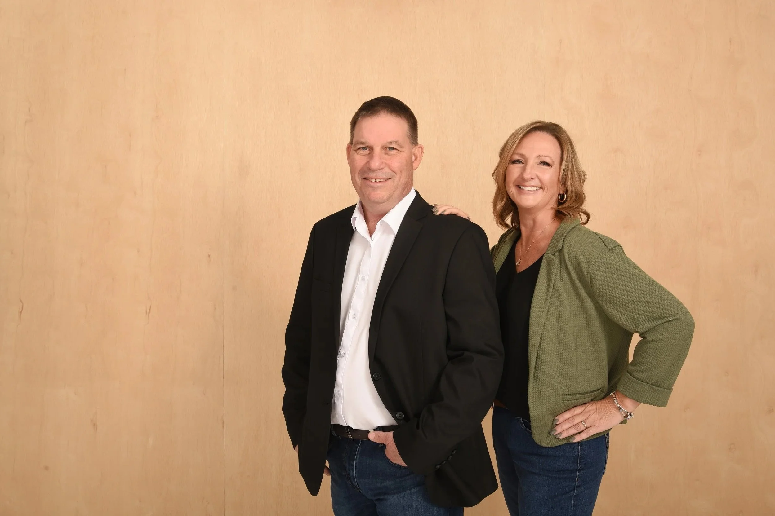 A man and woman standing together against a plain, light-colored wall. The man is wearing a black blazer over a white shirt, with his hands in his pockets and smiling. The woman is wearing a green jacket over a black top, with her right hand on her hip and smiling. She has her left hand resting on the man's shoulder.