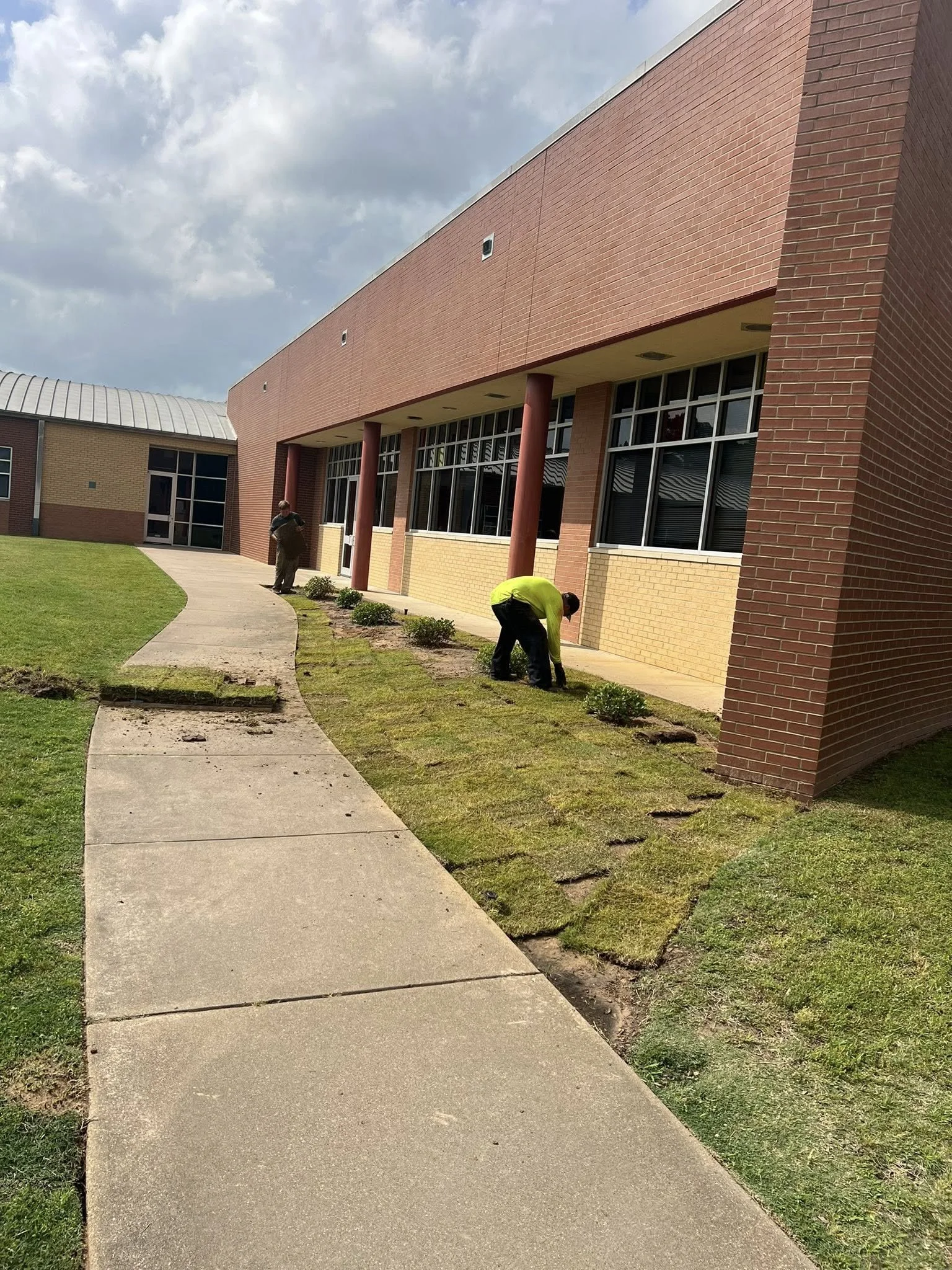 Two workers planting new grass and maintaining landscaping along a sidewalk outside a brick building under a partly cloudy sky.