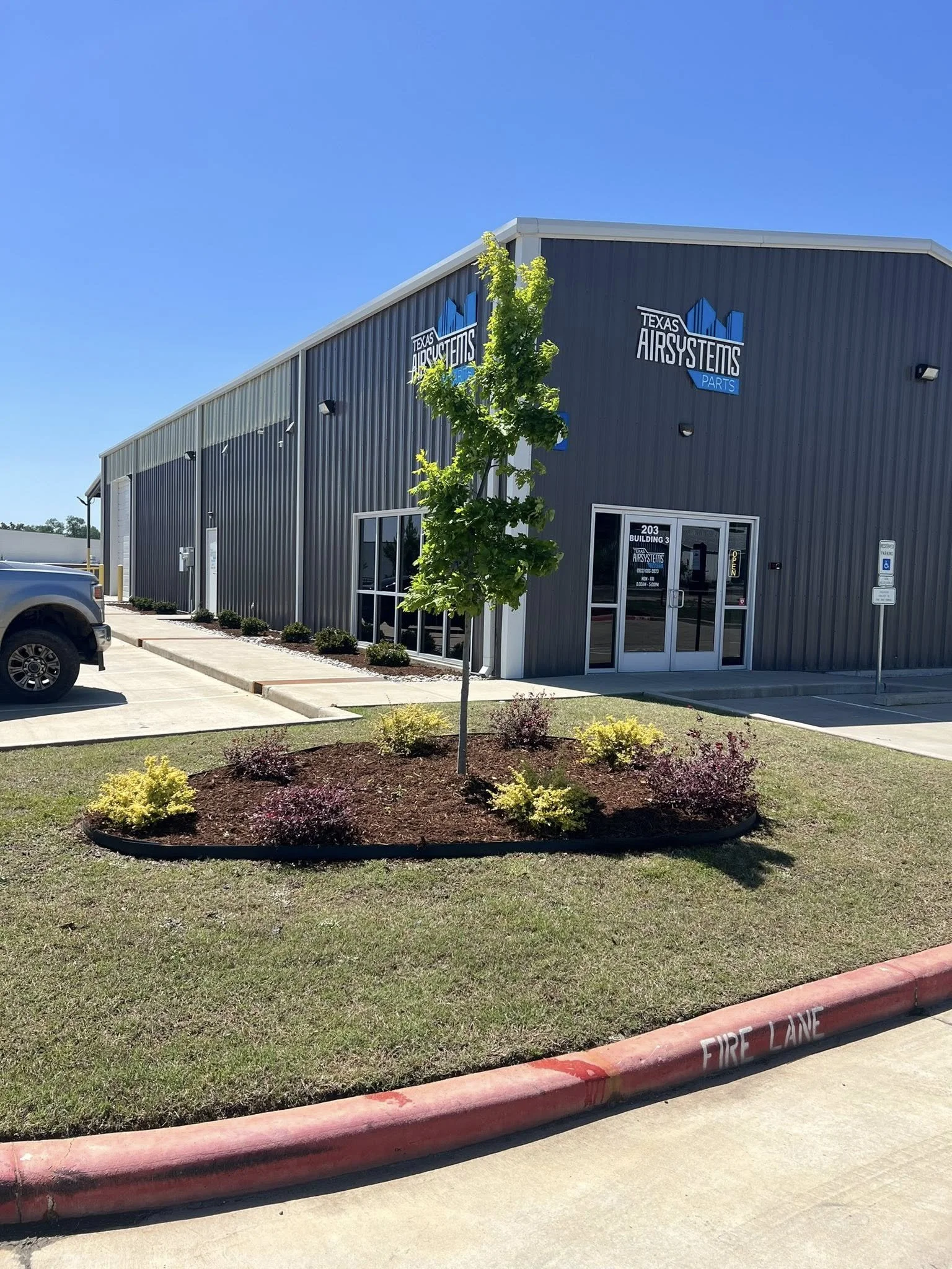 Exterior view of a metal building housing Texas Air Systems Parts, with a parking lot, a small landscaped area with a young tree and bushes, and a fire lane in the foreground on a clear, sunny day.