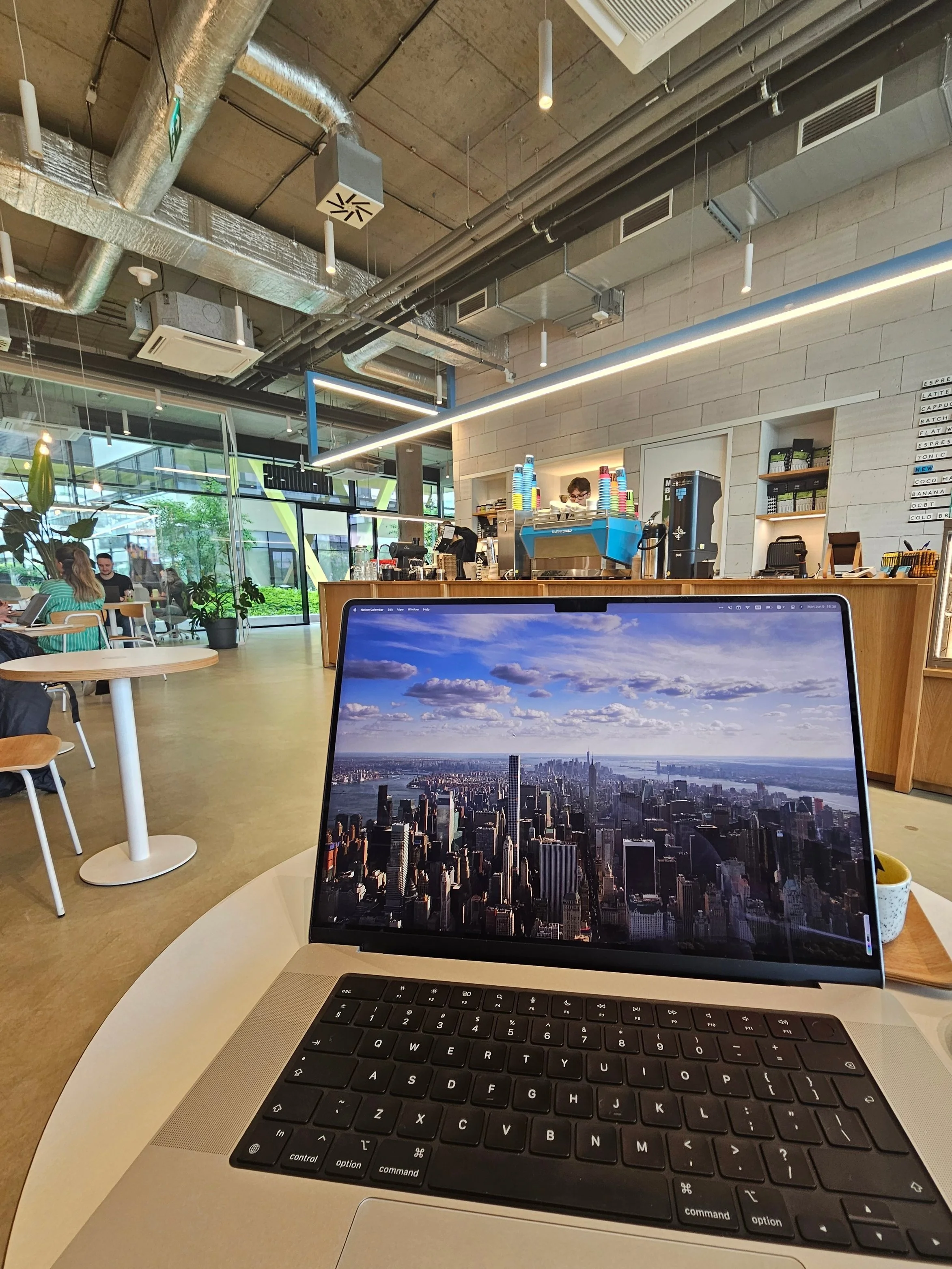 A laptop displaying a cityscape view of New York City, with tall buildings and partly cloudy sky, on a white round table in a modern coffee shop with wooden furniture and exposed industrial ceiling.