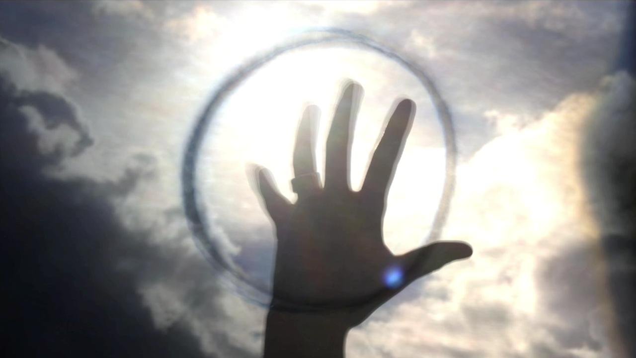 A person’s hand is silhouetted against a bright sky with cloud formations, reaching upward towards the sun, creating a halo effect around the hand.