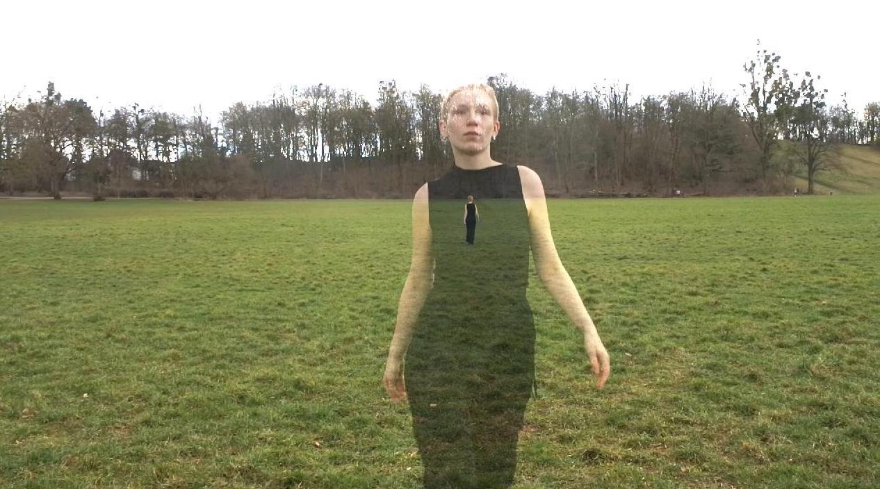 An overcast outdoor scene with a woman appearing semi-transparent, wearing a black sleeveless dress, walking on a grassy field with trees in the background.