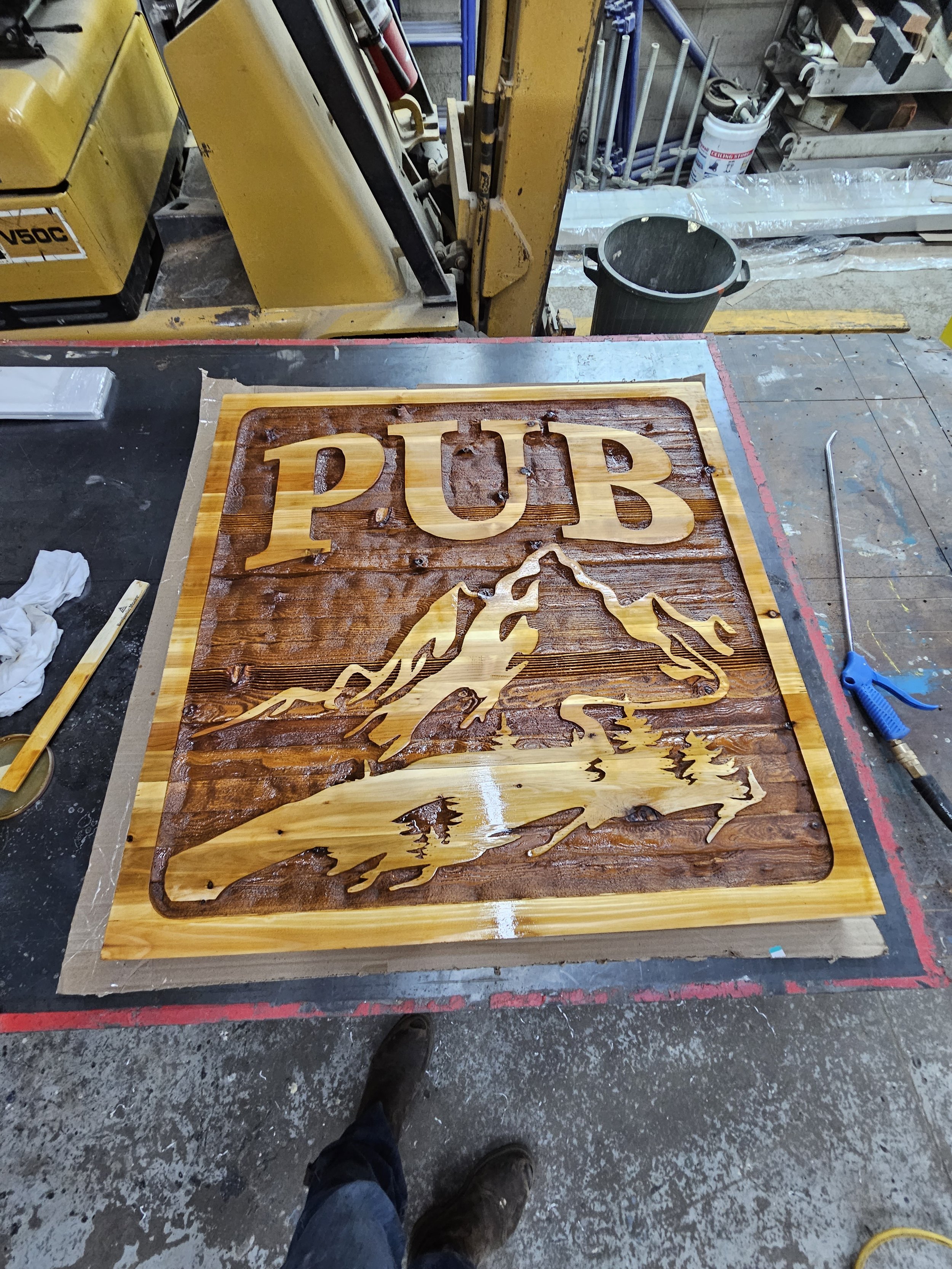 Wooden wall art featuring the word 'PUB' and a depiction of a mountain, with a river flowing at the base, in a workshop setting.