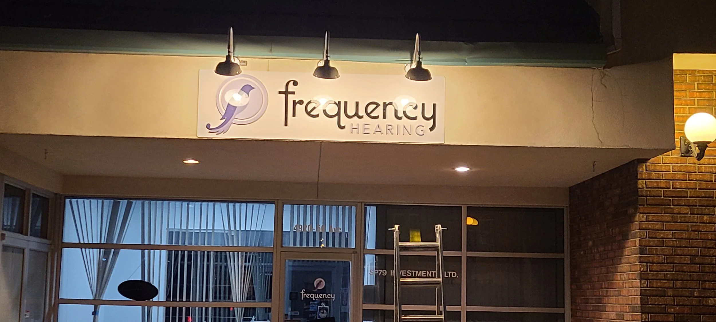 Sign for Frequency Hearing with a logo of a blue and white bird, outdoor wall-mounted lights, and a brick wall on the right side.