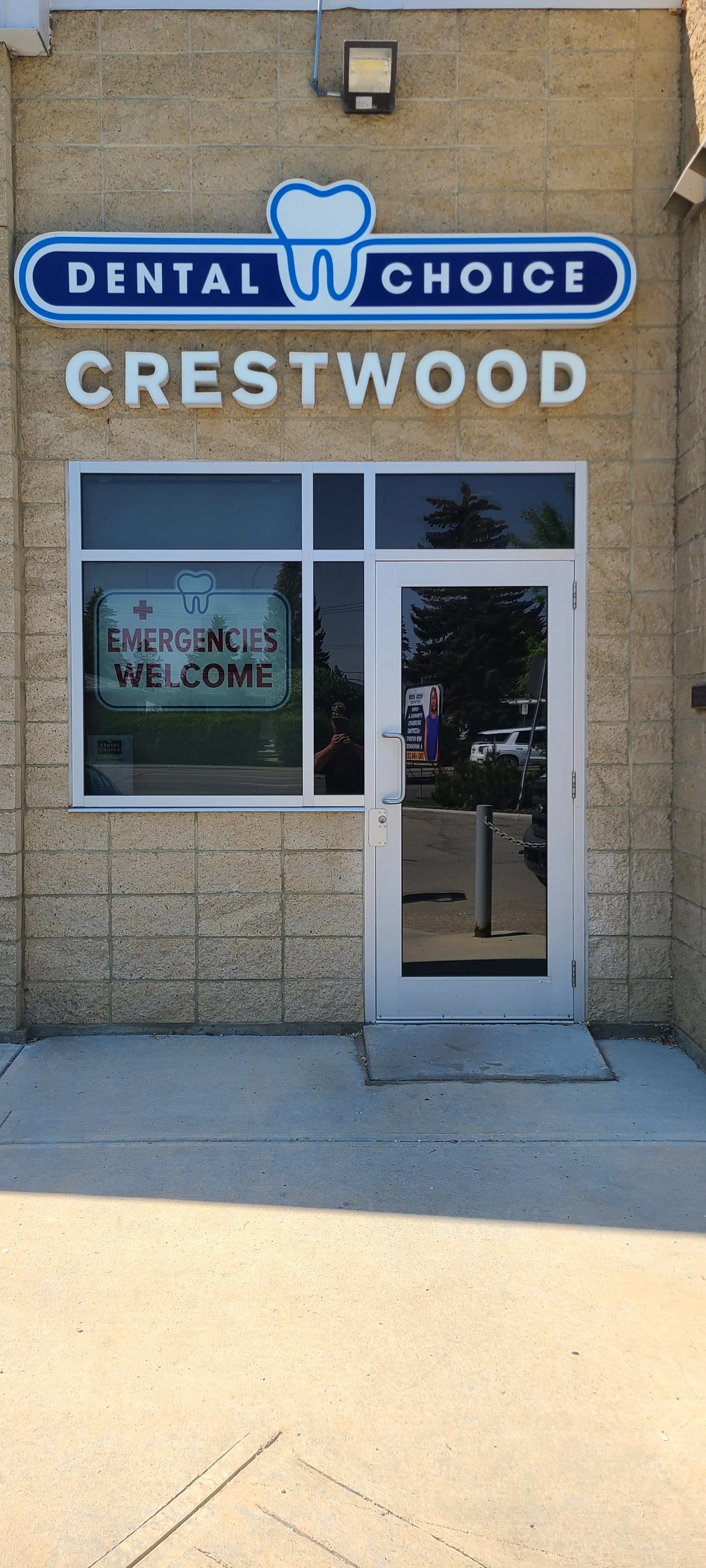 The exterior of a dental clinic named Crestwood with a sign that reads Dental Choice. The window has a sign that says Emergencies Welcome, and the door is made of glass with a metal handle.