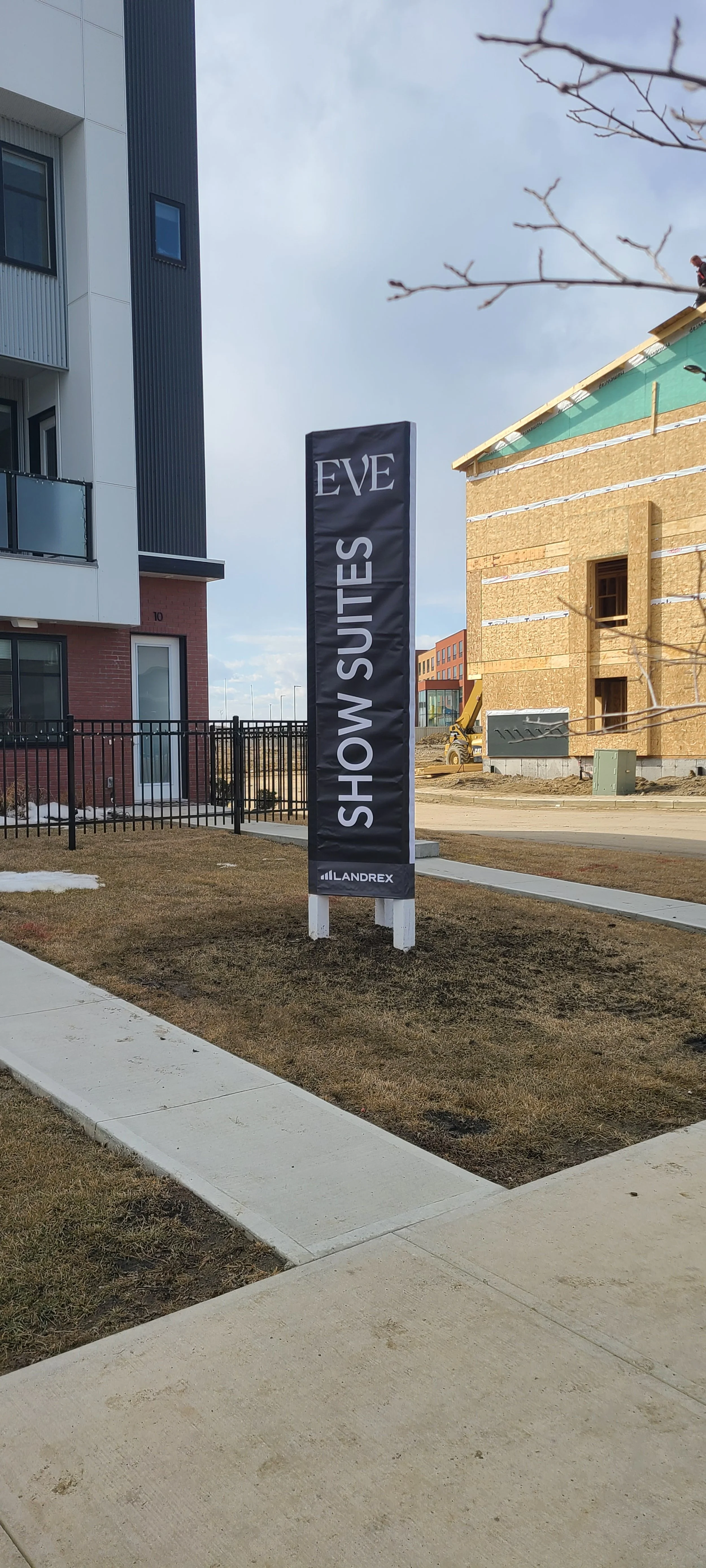 A black vertical sign with white lettering reads 'EVE SHOW SUITES' and is placed on a grassy patch outside near a sidewalk, with a modern apartment building and construction site in the background.