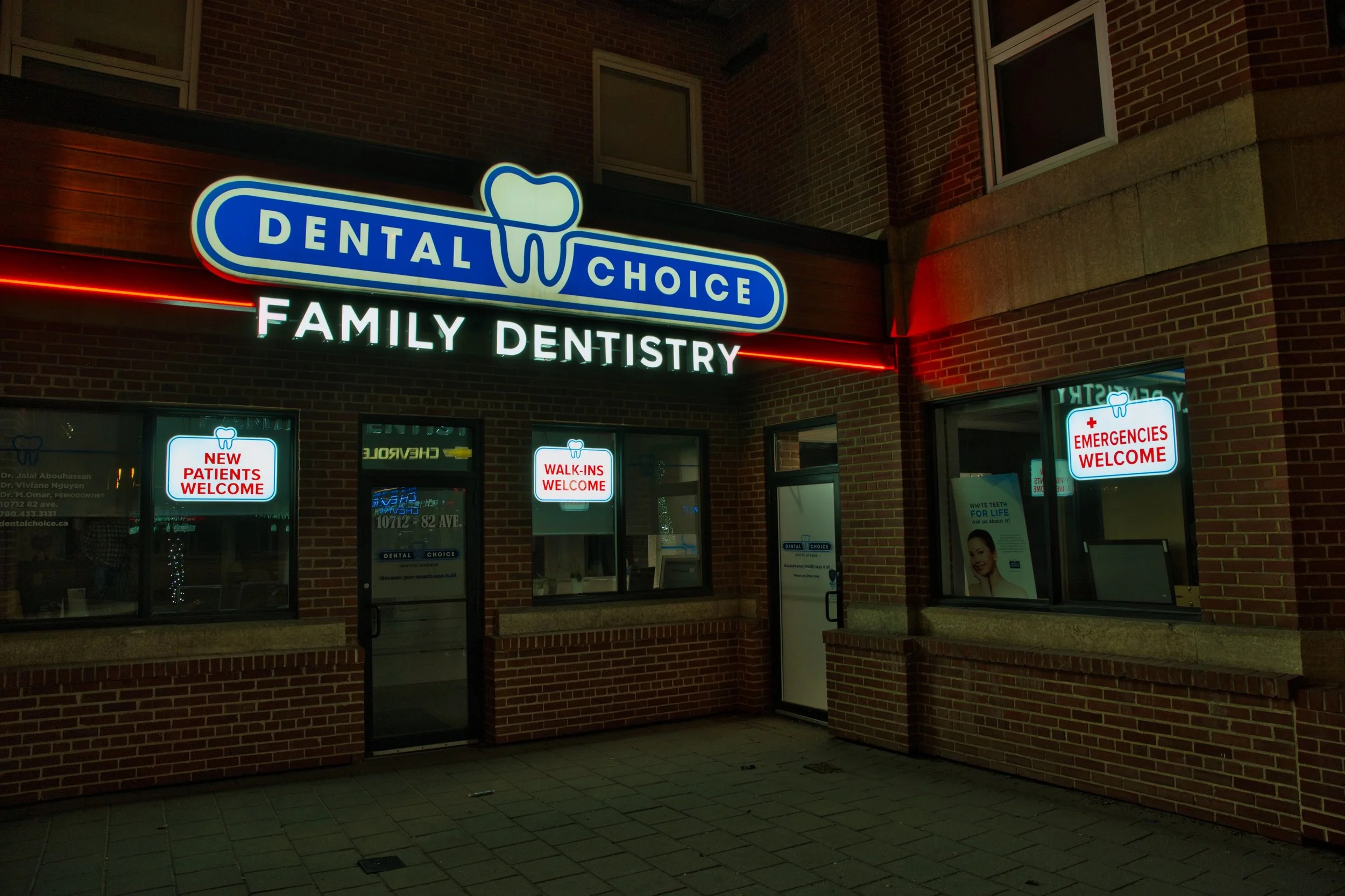 Night view of a brick dental clinic with illuminated signs reading Dental Choice Family Dentistry, with window signs that say 'New Patients Welcome,' 'Walk-ins Welcome,' and 'Emergencies Welcome.'