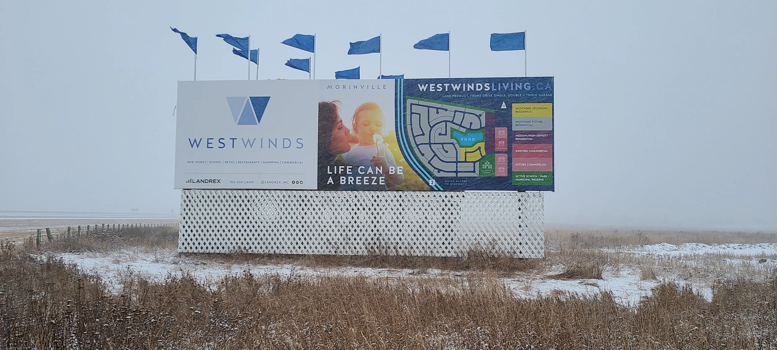Large billboard advertisement for Westwinds development, showing a map and promotional message, on a snowy field with a foggy sky and blue flags above.