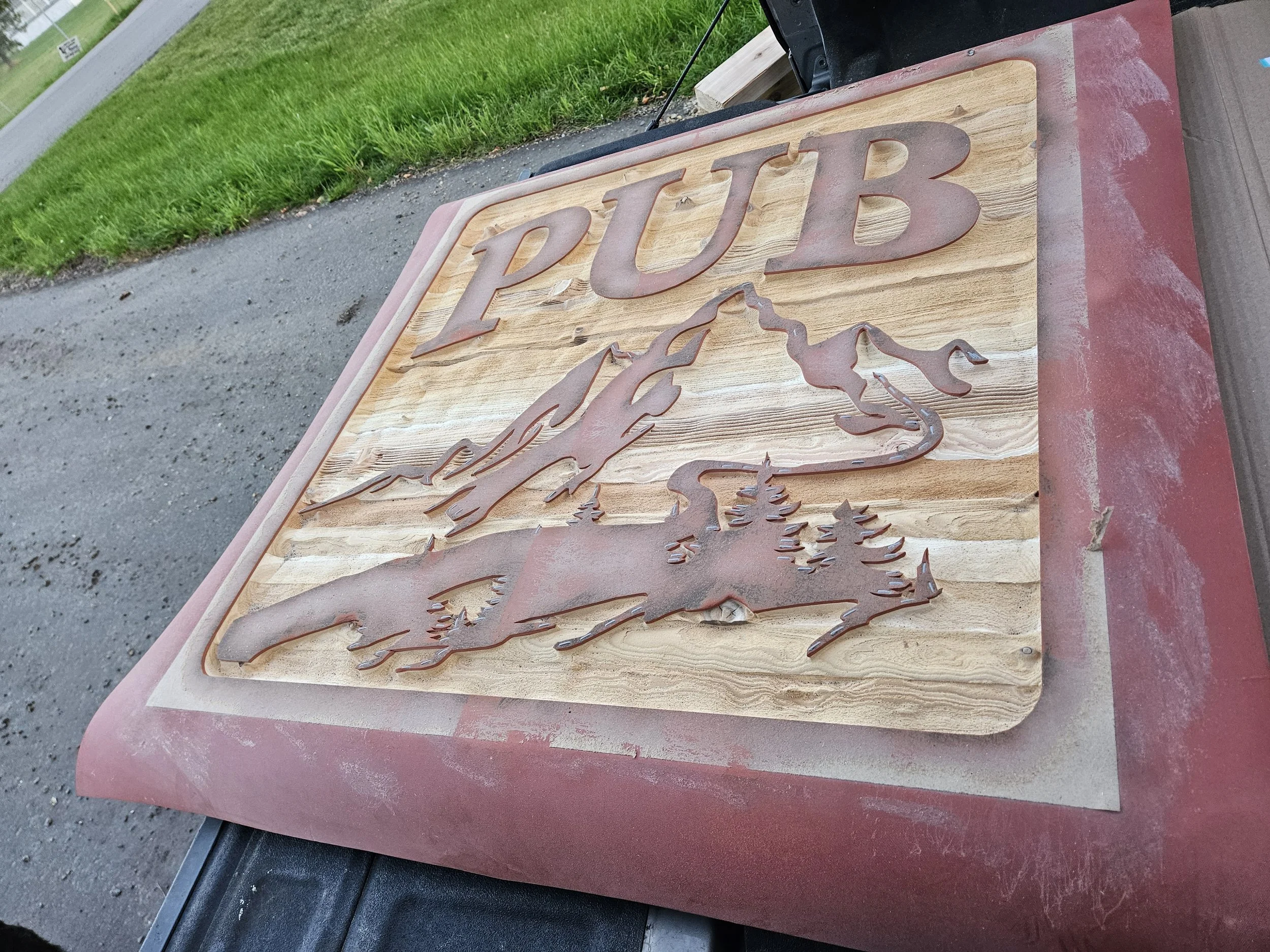 A wooden sign with the word 'PUB' and a mountain and tree scene cut out, resting on a red surface outdoors.