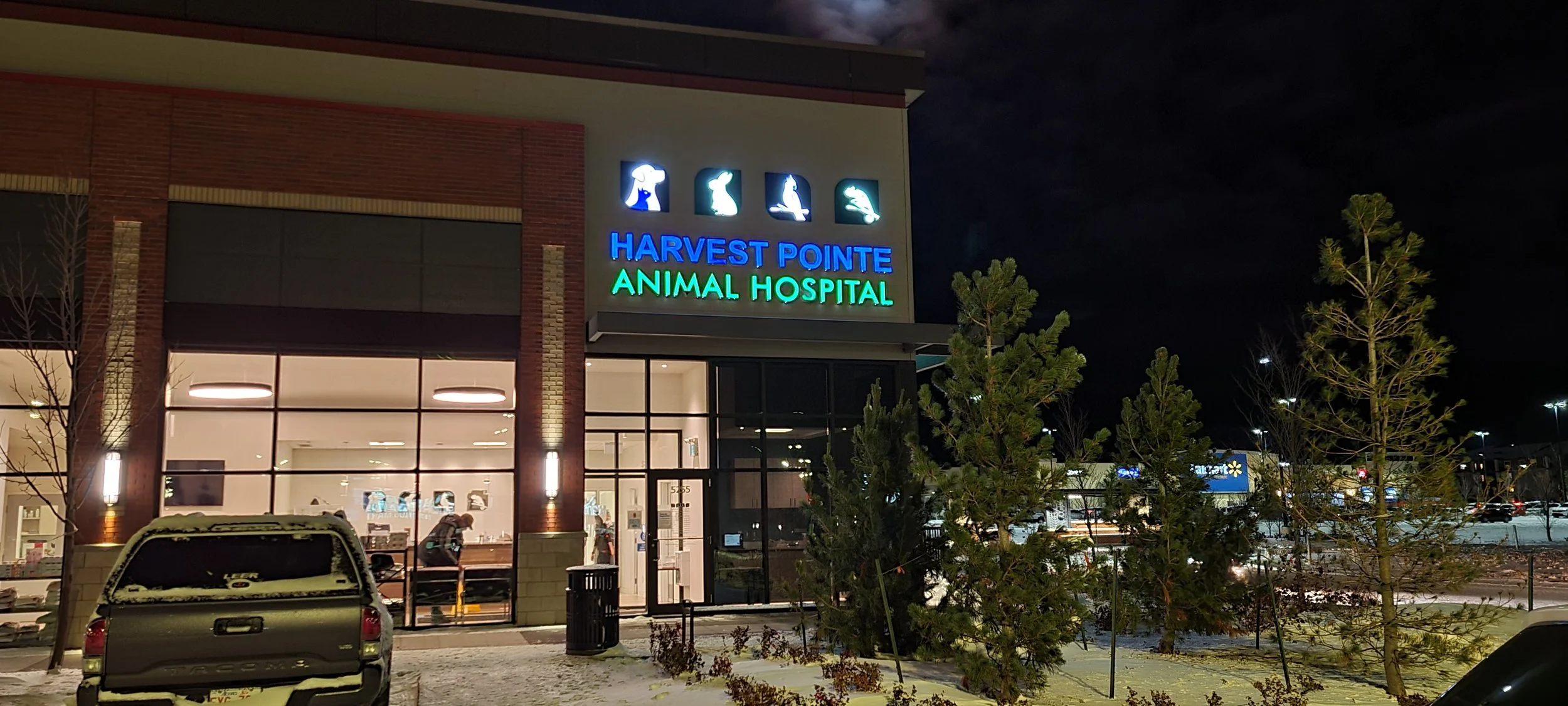 Night view of Harvest Pointe Animal Hospital with illuminated sign featuring icons of different animals and interior visible through glass doors.
