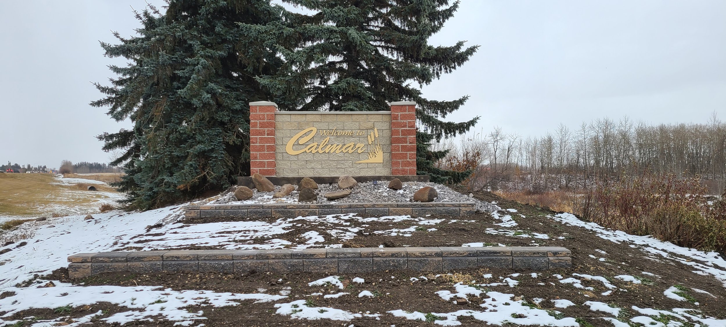 A welcome sign for Calmar, built with brick and stone, situated on a small landscaped mound with rocks, under snow, with a large evergreen tree behind it, and a background of leafless trees and a cloudy sky.