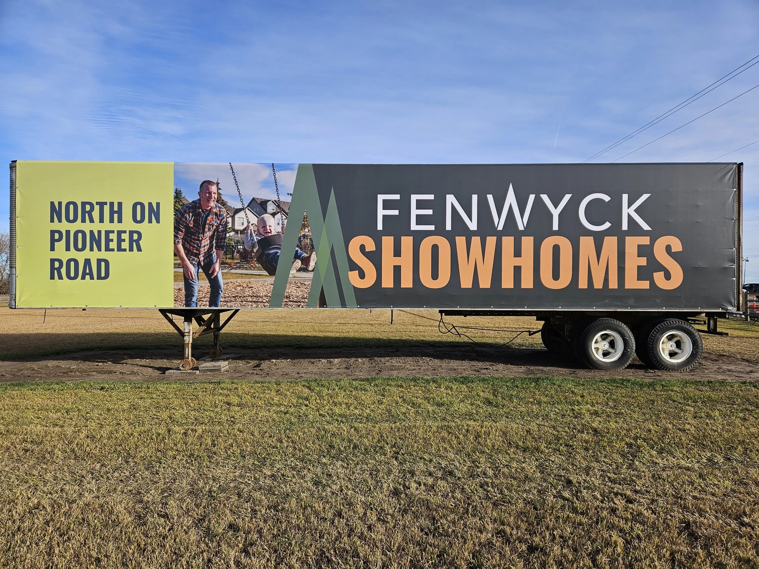 A banner advertising Fenwyck Showhomes on Pioneer Road, showing a man and a child on a swing set in front of a suburban neighborhood.