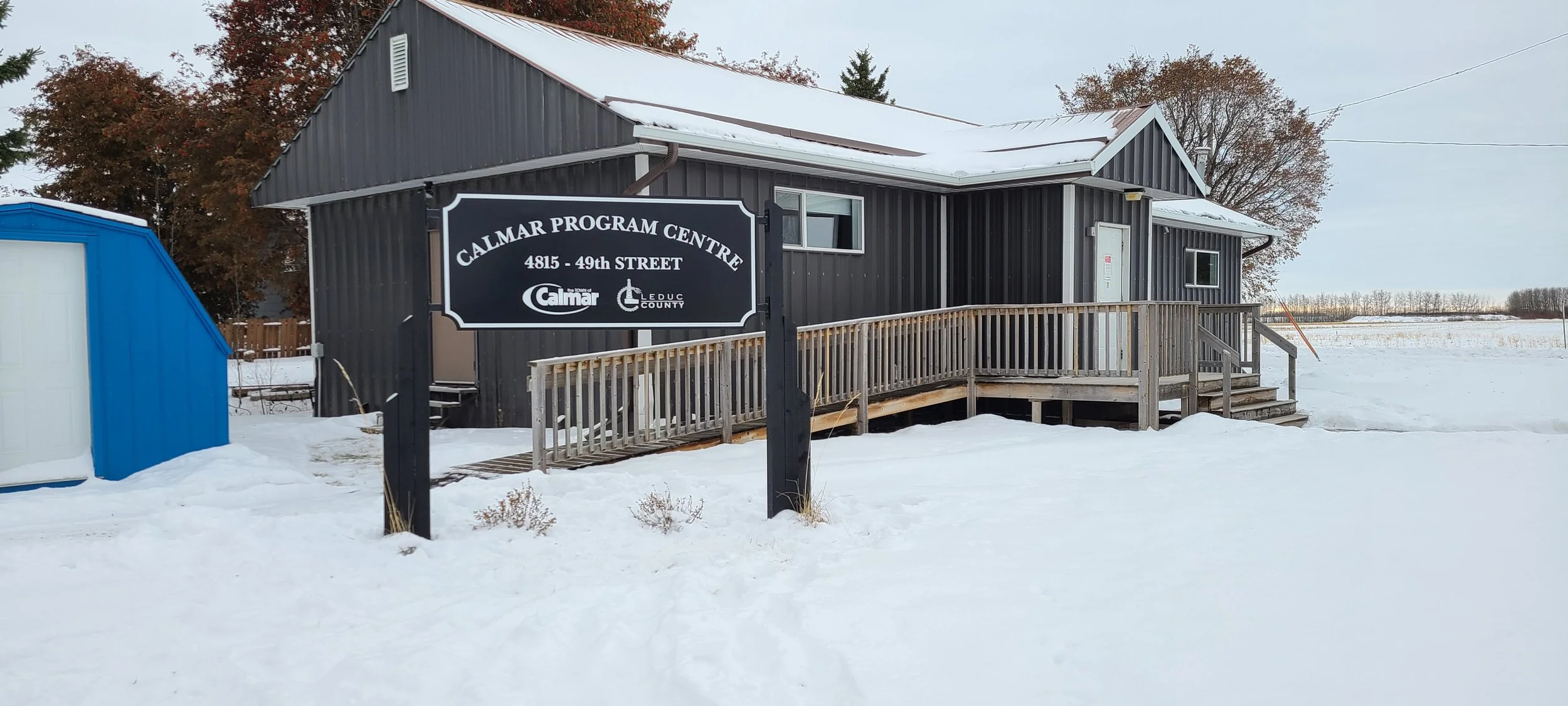 Black building with a sign reading 'Calmar Program Centre' on snow-covered ground, with a blue shed nearby and trees in the background.