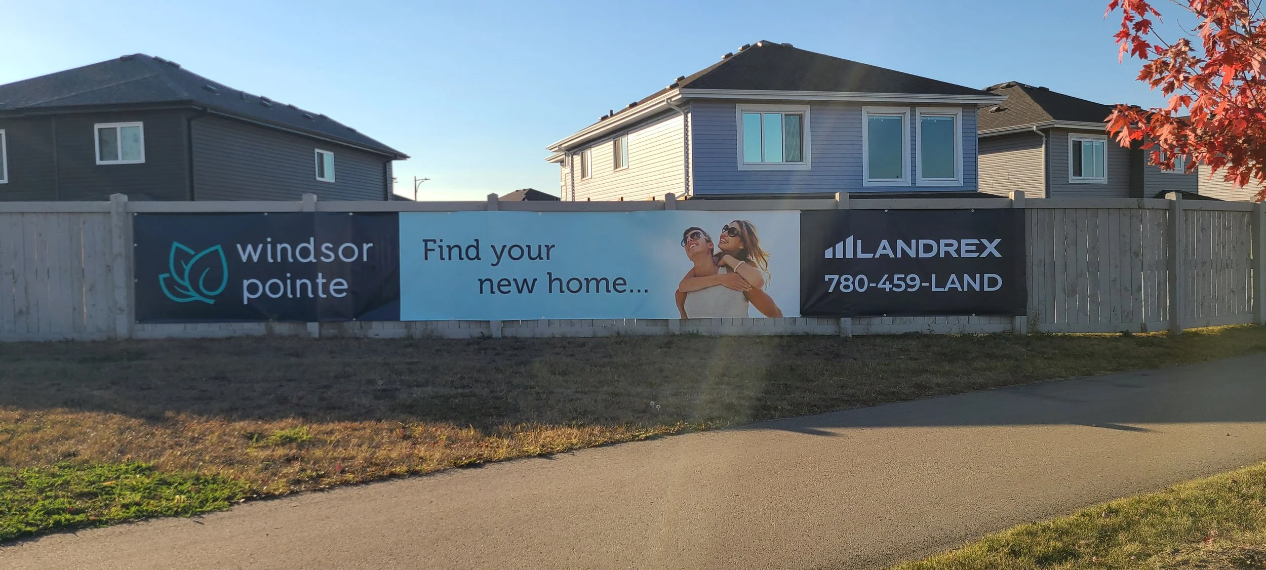 Real estate sign on a wooden fence in front of two blue houses reads 'Windsor Pointe, Find your new home...' with a logo and contact number '780-459-LAND'.