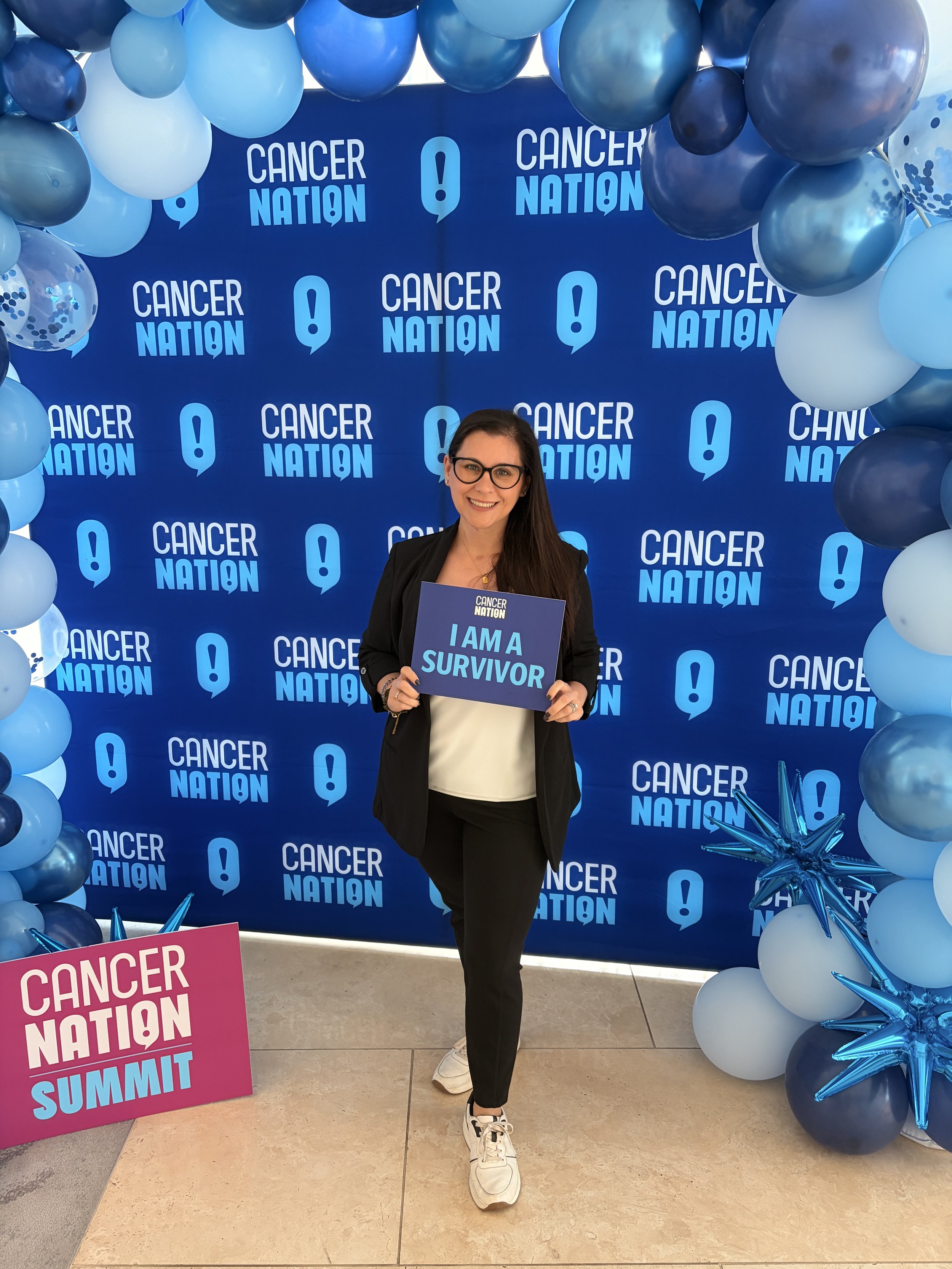 Tracy Steffek, founder of Remedy & Root, standing in front of a Cancer Nation banner holding a sign that says I'm a survivor.