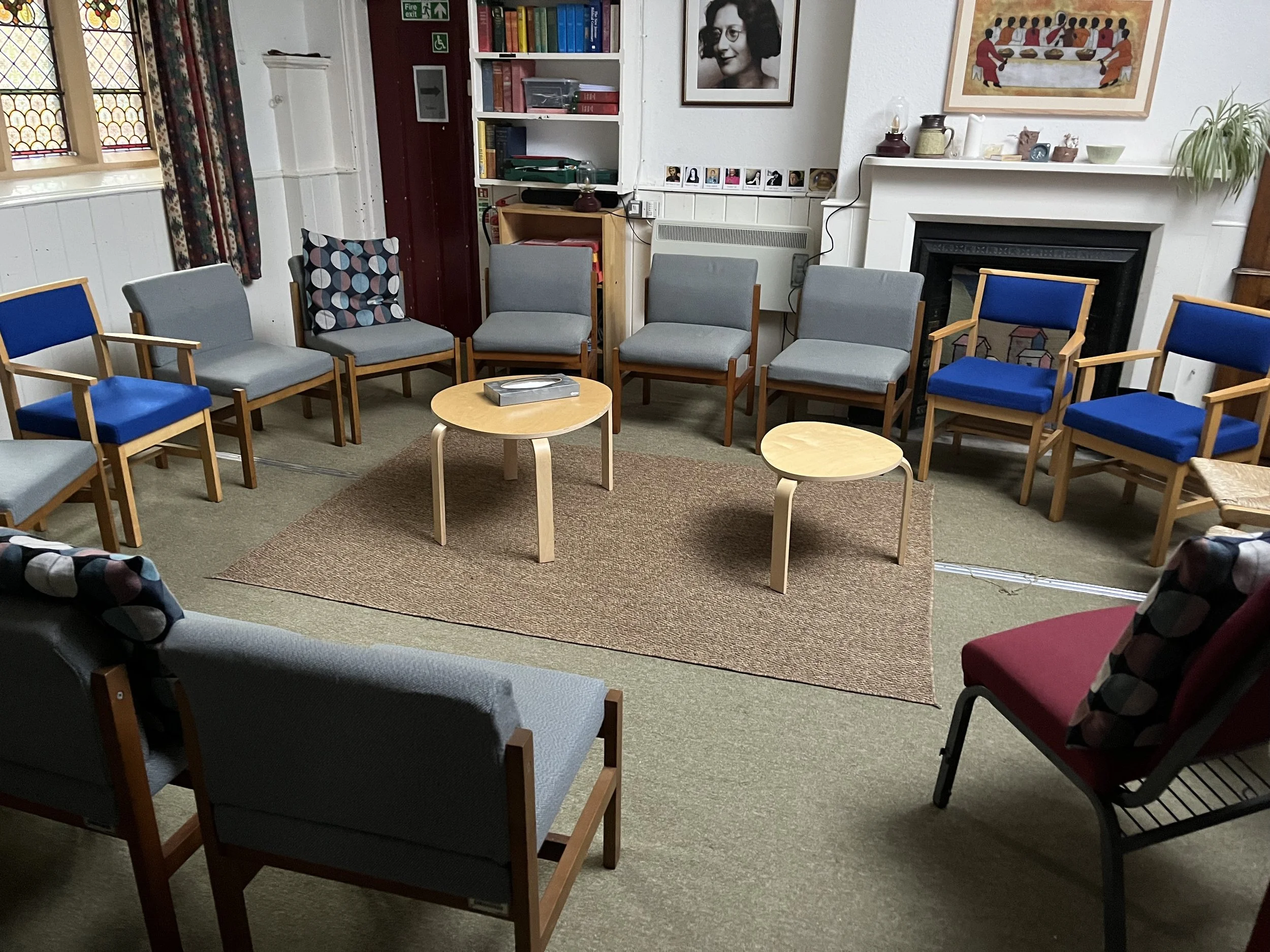A circle of chairs arranged in a room with beige carpet, a small oval coffee table and a rectangular table, in front of a fireplace with decor and wall art, and a bookshelf with books and photos.