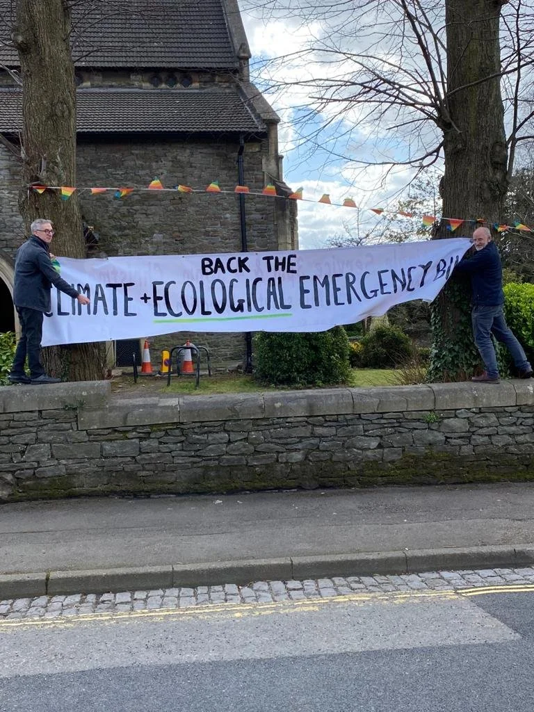 Two men are holding a large protest banner that reads 'BACK THE CLIMATE + ECOLOGICAL EMERGENCY' in front of an old stone building with trees and colorful bunting hanging above.